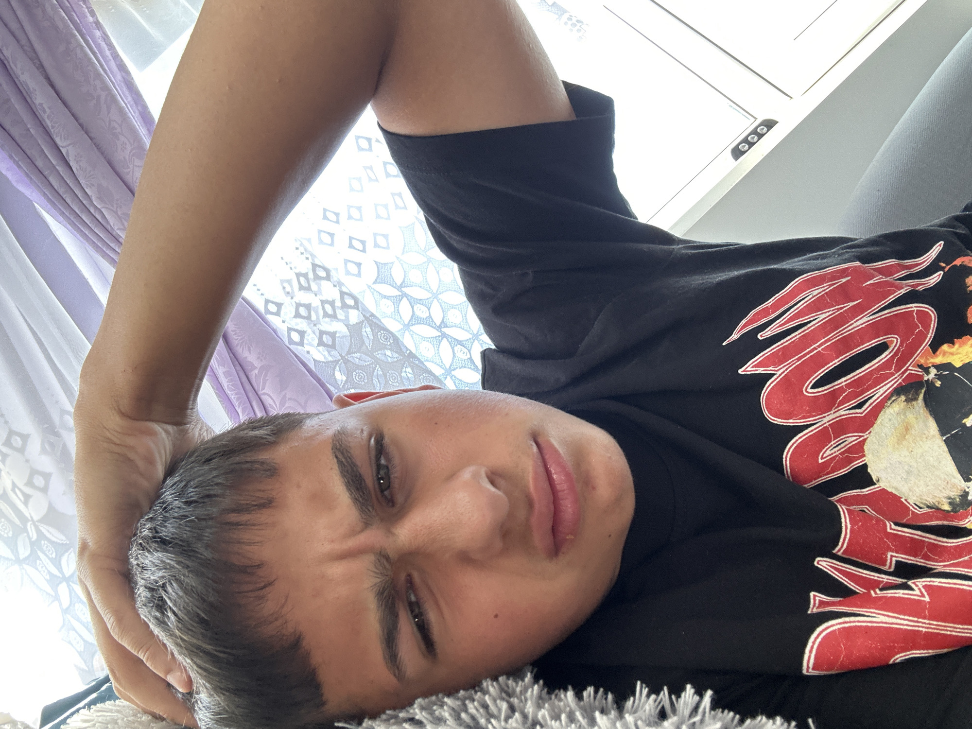 A young male is in a close-up, selfie-style shot, indoors. He has short, dark hair and is wearing a black t-shirt with a red graphic design featuring flames and lettering. His right hand is resting on the top of his head. His facial expression is a grimace, with his eyebrows furrowed and his lips pursed. The background consists of draped fabric in light purple and white, with a patterned sheer curtain to the right. A light-colored, textured object, possibly a blanket or pillow, is visible on the left side of the frame. The lighting suggests it is daytime, with light coming from the right.