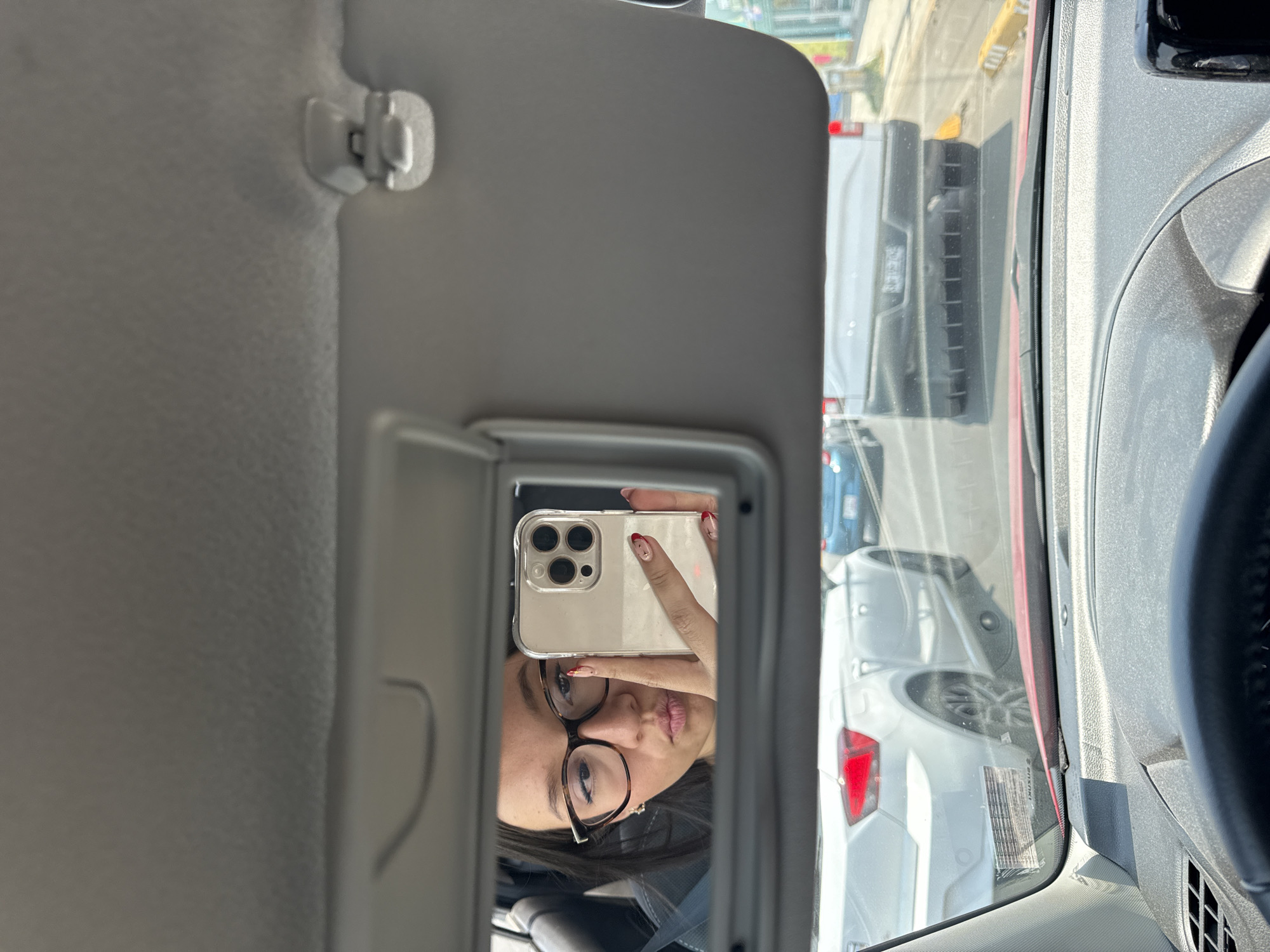 The image is taken from inside a car, capturing the reflection in the sun visor mirror. The reflection shows a woman with glasses, holding a phone up to take a selfie. Outside the car window, the image reveals a street scene with other vehicles, suggesting traffic. The location is identified as San Bernardino Tlaxcalancingo, Mexico. The composition includes interior elements of the car, such as the dashboard and steering wheel, alongside the exterior environment visible through the window and reflected in the mirror.