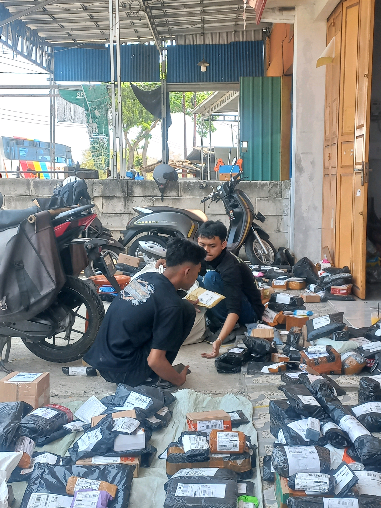 The image depicts an outdoor scene, likely a small business or distribution point, in Baturetno, Indonesia. Two young men, wearing dark t-shirts and casual pants, are engaged in sorting and organizing numerous packages. The packages vary in size and packaging, with many enclosed in black plastic or brown cardboard. Some packages have shipping labels affixed.
The environment appears to be a covered outdoor area with a metal roof supported by pillars. To the right, there are wooden doors of a building. In the background, a bus with colorful stripes is visible, suggesting proximity to a road or transportation hub. Two motorcycles are parked nearby, one with a dark brown saddlebag. The ground is a mixture of concrete and a patterned textile or tarp, upon which the packages are spread. Trees and some foliage are visible in the distance, indicating a semi-urban or rural setting. The lighting suggests it is daytime, with sunlight casting shadows. The overall impression is one of active logistics and package handling.
