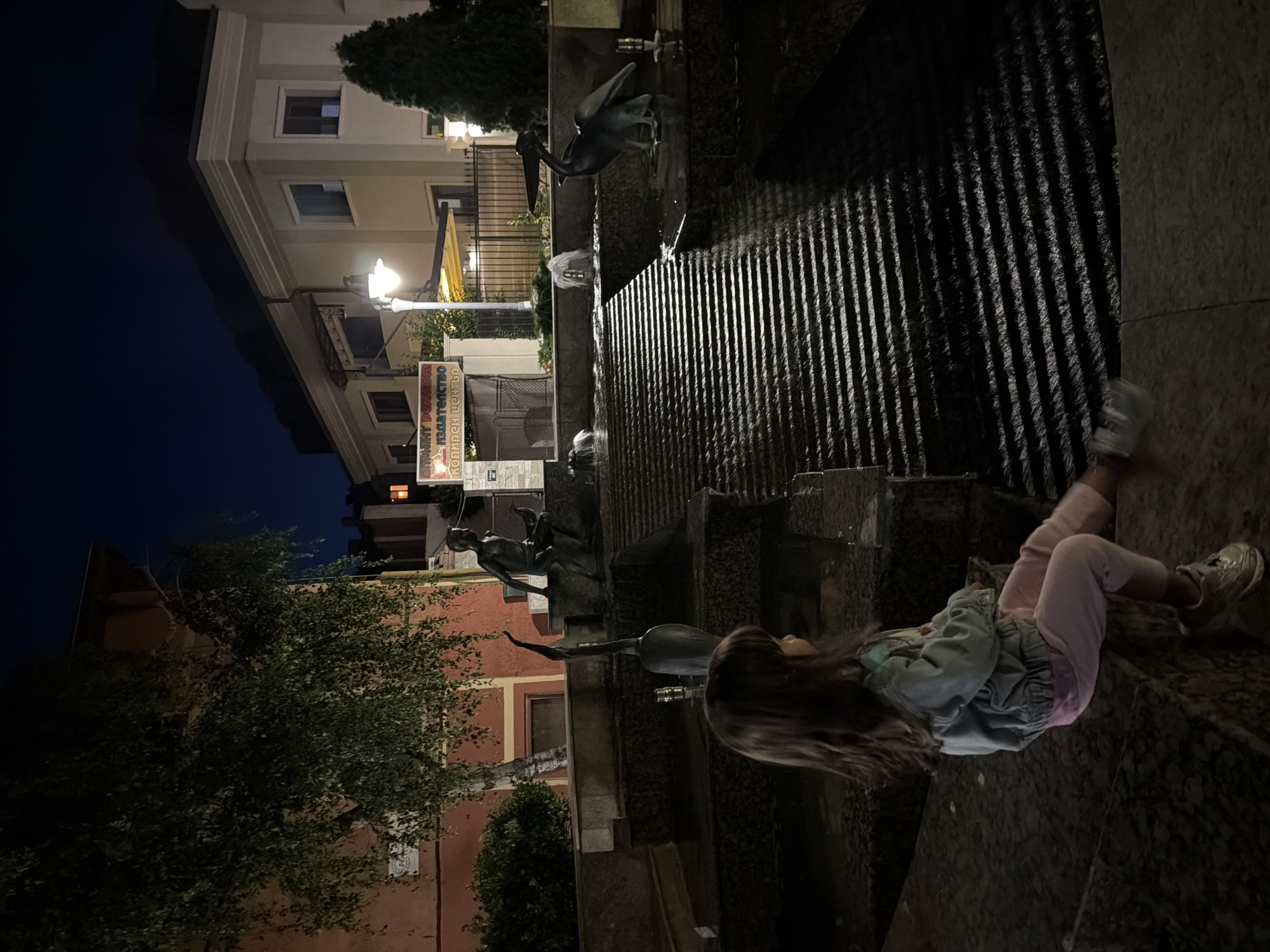 A young girl with long brown hair sits on the edge of a stone fountain at night in the city of Burgas, Bulgaria. She is wearing a light blue denim jacket over a pink shirt and pink pants. Her feet are visible, she is wearing silver sneakers and is looking to the right. The fountain has multiple tiers with water flowing down in horizontal lines. Two bird statues, one resembling a crane and another a pelican, are integrated into the fountain's design. In the background, there are buildings and streetlights illuminating the scene. A statue of a young boy sitting on a ledge is also visible. A sign with Cyrillic text is posted on a building in the background. The sky is dark blue, indicating it is nighttime.