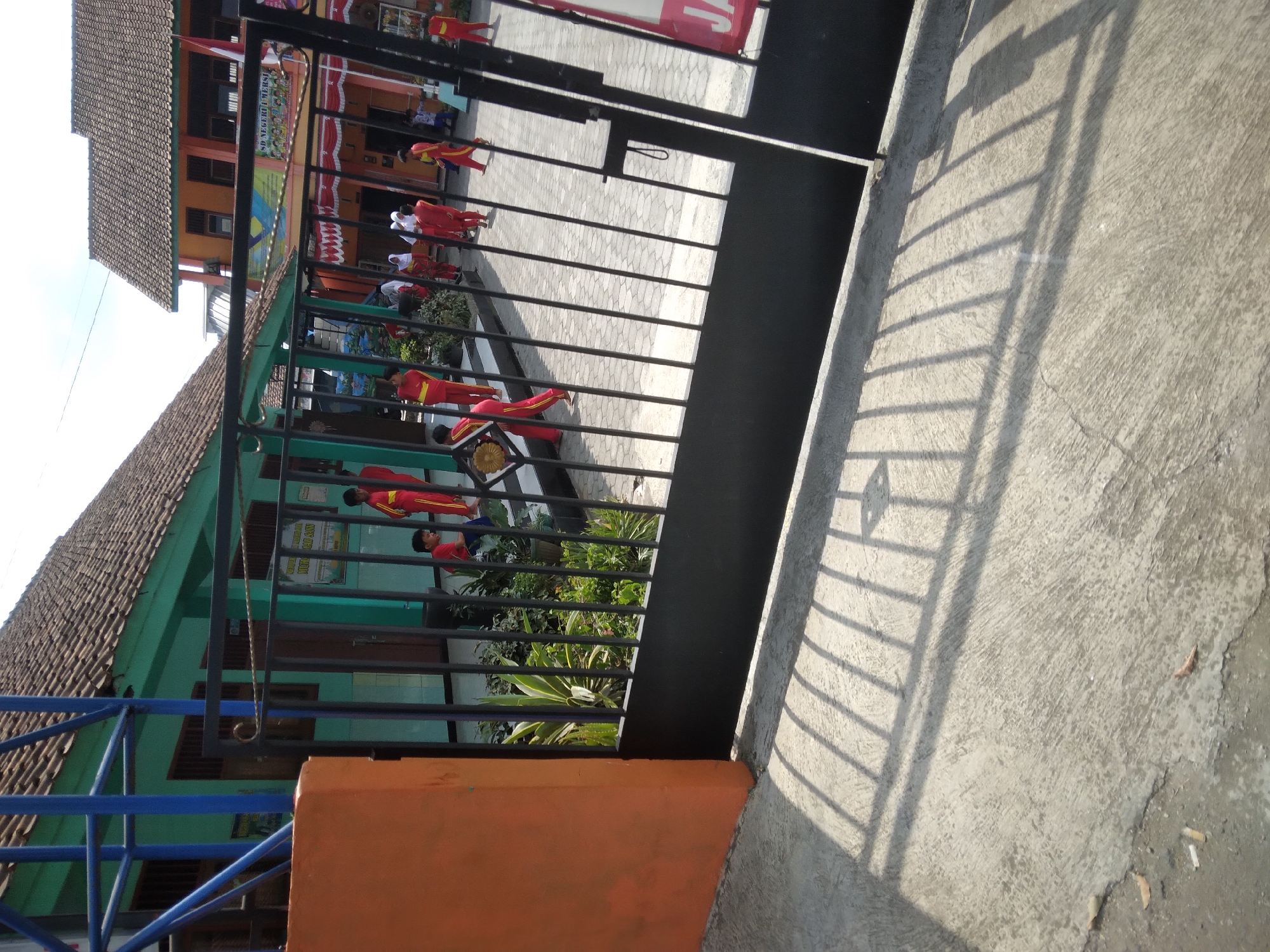 The image shows an outdoor view of a school in Sokaraja, Indonesia, likely during the daytime. A black metal gate is in the foreground, casting long shadows on a concrete walkway. Beyond the gate, several children in red and yellow uniforms are visible on a paved courtyard. They appear to be students, some walking and others standing. In the background are two school buildings with tiled roofs. The building on the left is painted green, and the one on the right is orange. There are also signs and flags visible on the buildings. Plants and greenery are present in front of the green building. The overall scene depicts a typical school environment.