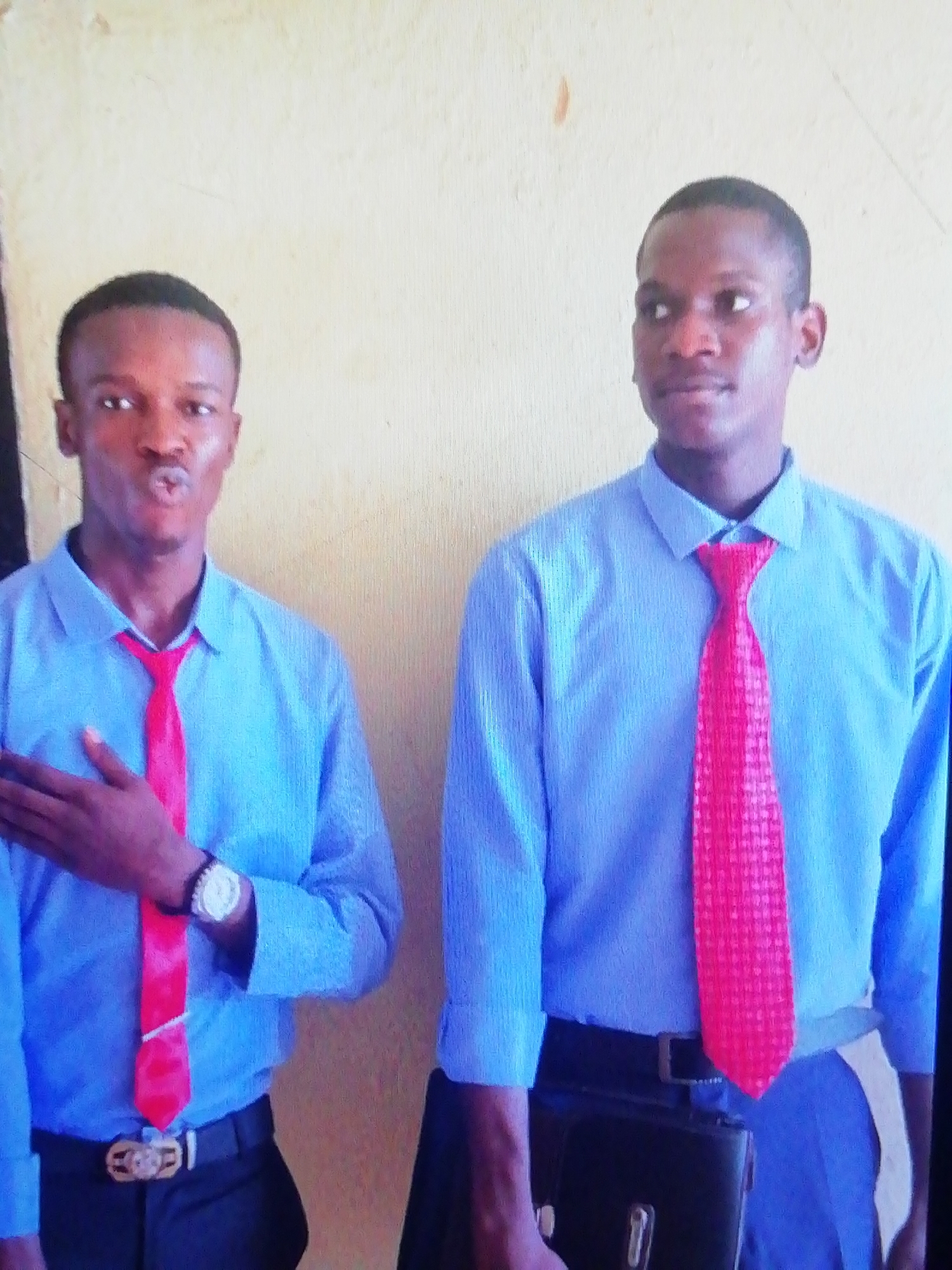 Two young men are standing indoors against a light-colored wall. Both are wearing light blue, long-sleeved shirts and red ties. The man on the left has his hand over his heart and is puckering his lips. He is wearing a wristwatch and a belt with a large buckle. The man on the right is carrying a dark-colored briefcase. The location is possibly Enugu, Nigeria.