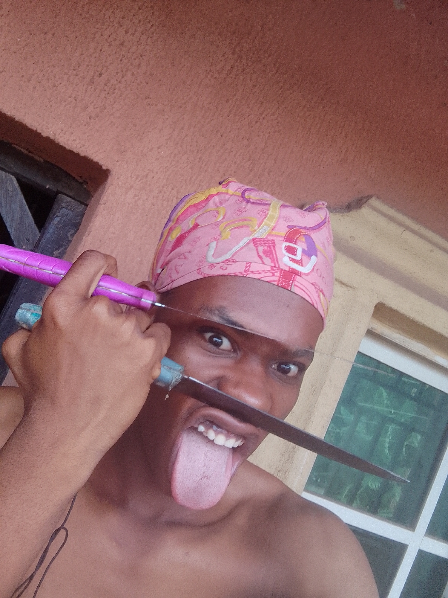 A shirtless person with a pink patterned head covering is holding two knives to their face, with one knife above the eyes and the other between the nose and mouth, sticking their tongue out. The knives have purple and blue handles. The background includes a textured orange wall, a dark wooden window frame, and a window with a white frame and green tint. Location is Unknown, Unknown.