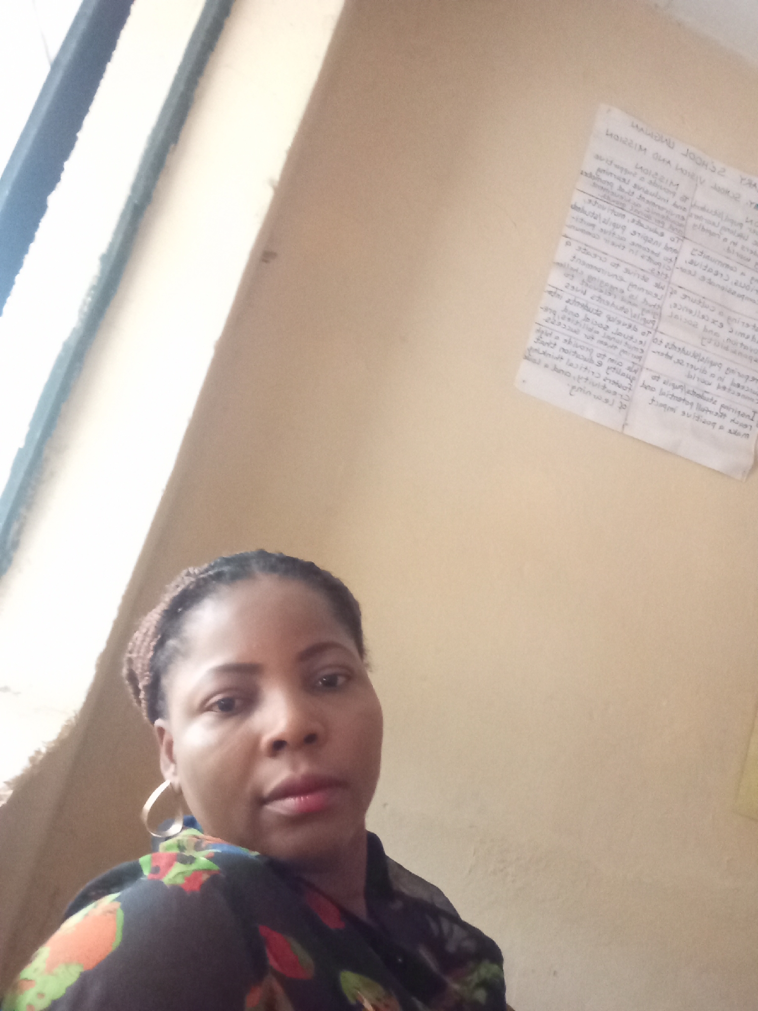 A medium shot captures a woman indoors. She has dark skin, braided hair, and gold hoop earrings. She wears a dark blouse with a floral pattern. Her gaze is directed towards the camera. Behind her, a wall is visible, with a piece of paper attached. A window frame with what looks to be a white or light-colored panel is seen in the upper left corner. The city and exact location are not discernable from the image.