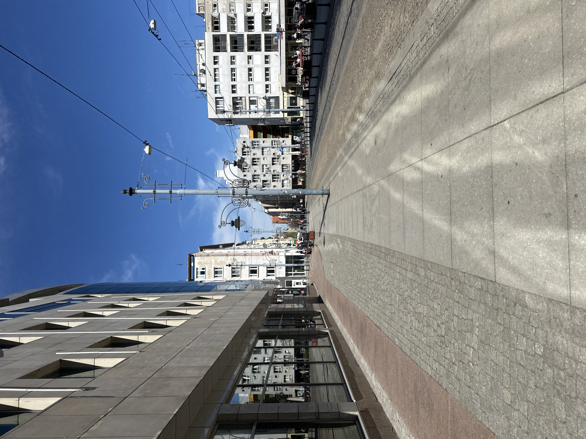 The photograph depicts a street in Wrocław, Poland, characterized by modern and older architectural styles. The image is taken at street level, looking down a thoroughfare. A modern, multi-story building with a textured, gray facade and distinctive window designs occupies the left side of the frame. The windows reflect the buildings opposite. On the right side, the street is lined with taller, older buildings with white facades and numerous windows. The street is paved with gray concrete slabs and a section of cobblestones with a reddish border. A decorative street lamp stands along the street. Overhead power lines crisscross the blue sky. People are seen walking and sitting at outdoor cafes along the street.