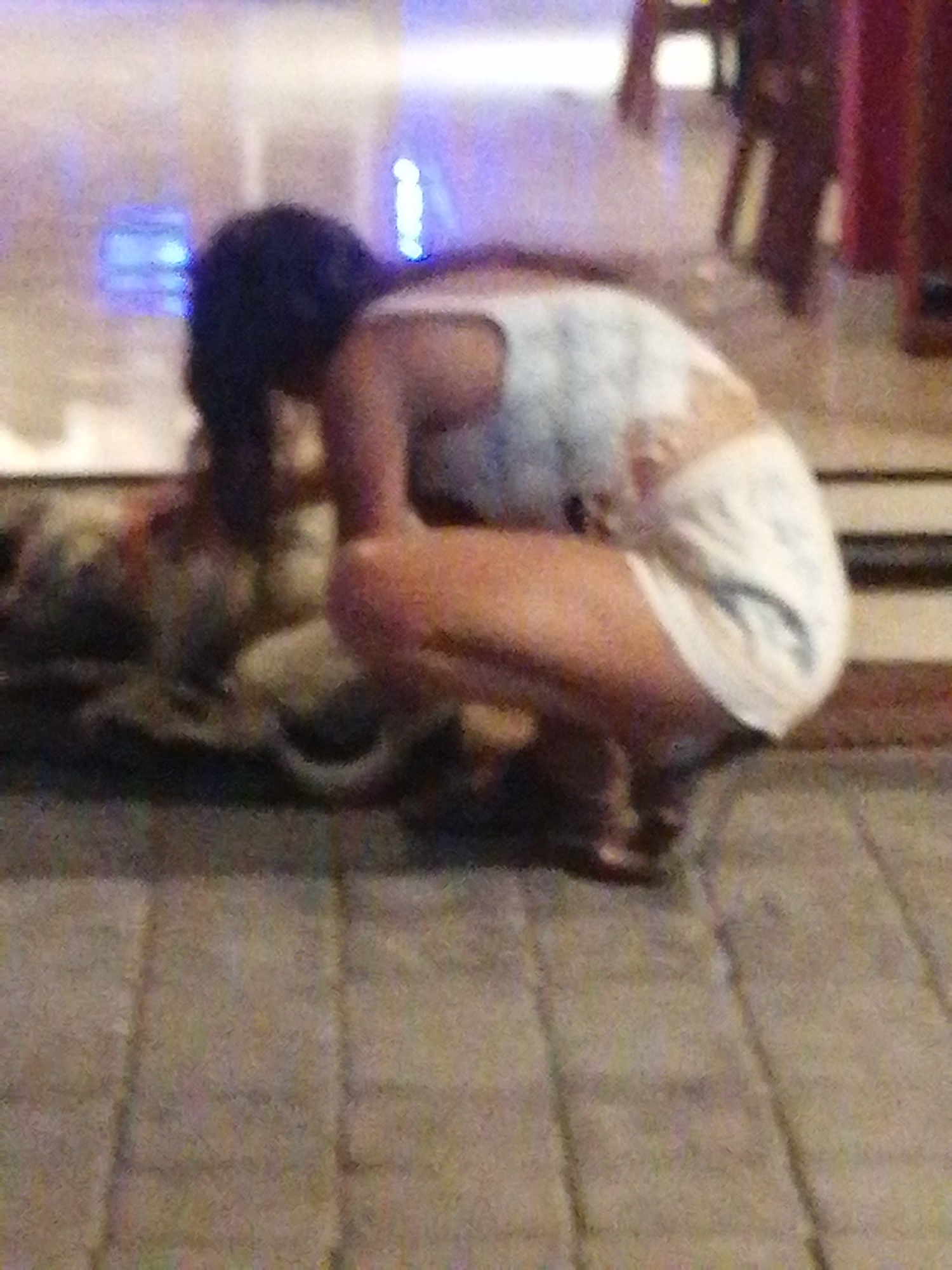 A person is crouched down on a cobblestone street in front of a blurred background that appears to be an indoor setting with lights. The person is wearing a white patterned top and white shorts. Their hair is dark and appears to be pulled back. There is a dog or animal partially visible next to the person. The scene suggests a casual encounter on a street, possibly at night due to the lighting in the background.