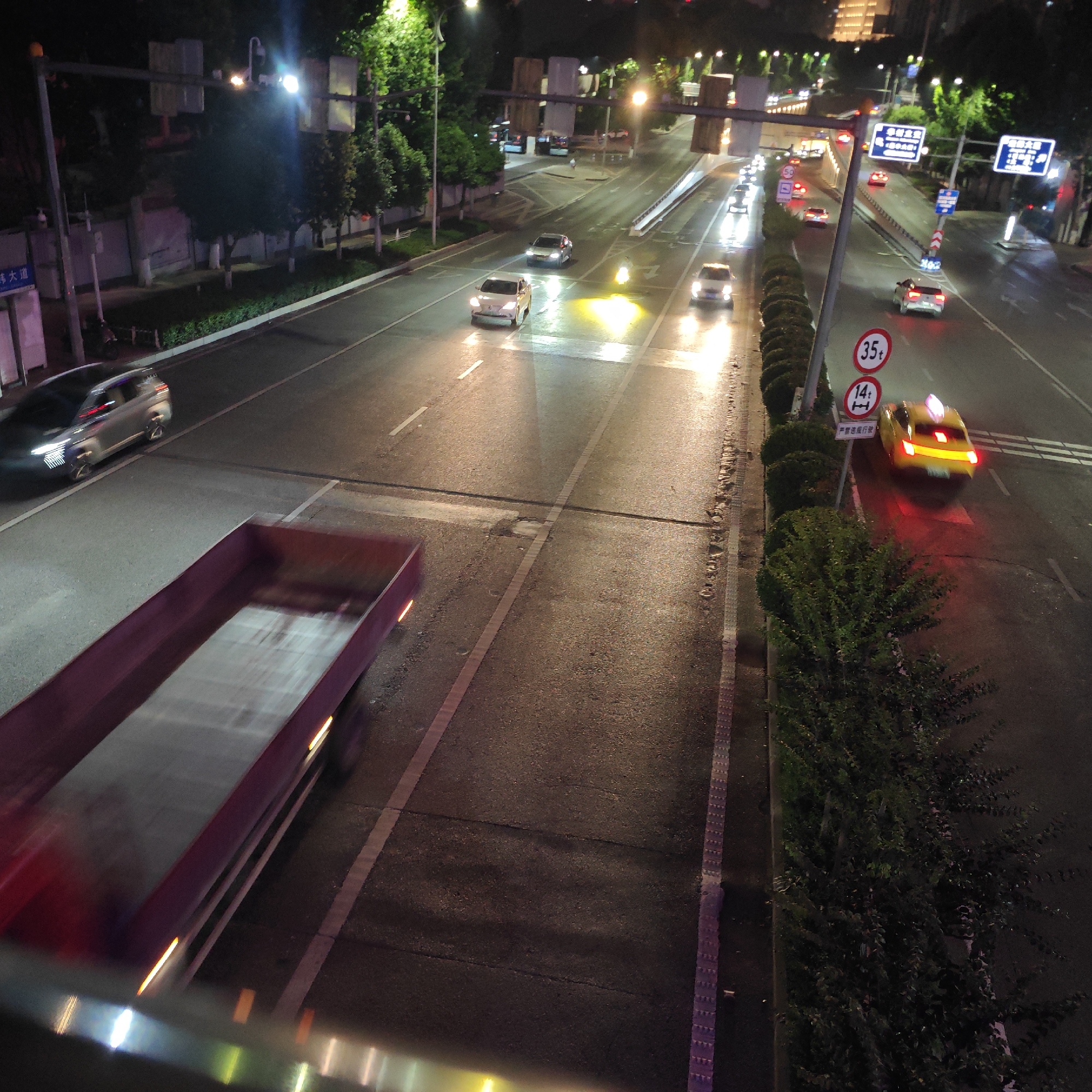The image depicts a night scene on a multi-lane road in the Yuzhong District of Chongqing, China. The road surface is wet, reflecting the headlights of the vehicles. A large truck is blurred in motion, traveling on the left side of the frame. Several cars are visible on the road, with lights illuminated. One appears to be a taxi due to the lit sign on top. Traffic signs and speed limit signs are visible along the right side of the road. A concrete barrier and some bushes line the right edge of the road. Streetlights illuminate the scene. The overall scene suggests low traffic at night.