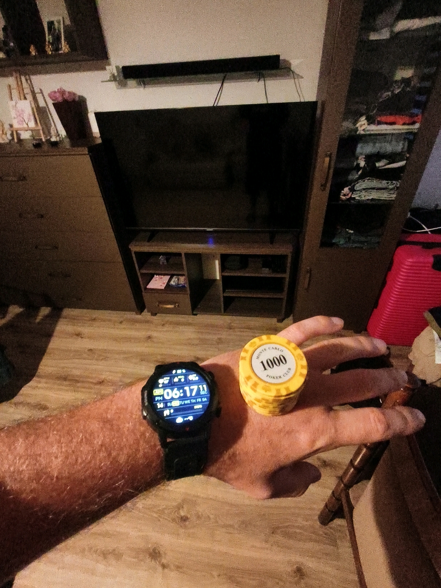 The image shows a close-up view of a person's left arm and hand, with a smartwatch on the wrist and a poker chip held in the hand. The smartwatch displays the time as 06:17. The poker chip is yellow and has "MONTE CARLO 1000 POKER CLUB" printed on it. The background suggests an indoor setting with furniture, including a television, a cabinet, and a suitcase. The lighting is dim. The location is described as Chojnice, Poland.
