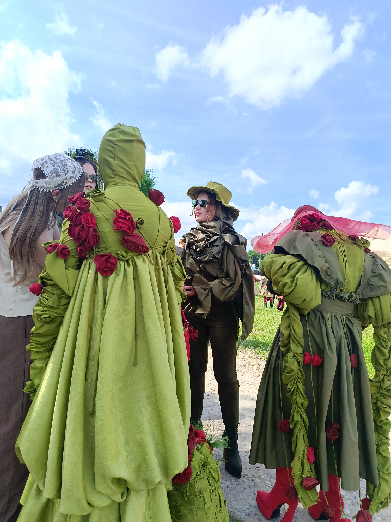 In Поповка, Russia, under a bright blue sky with scattered clouds, four individuals are captured in a medium shot. Three individuals are prominently featured in the foreground, two of whom are wearing elaborate, light green, hooded outfits adorned with red fabric roses. The outfit on the left has a voluminous skirt with ruched fabric. The third individual in the foreground, positioned between the two in green, wears a khaki-colored hooded outfit with a hat and sunglasses. Behind these three, a fourth individual in a large, elaborate pink and green hat with red roses is visible. The background shows a grassy field with some scattered structures and more people in the distance. The overall impression is a staged event or gathering with participants in distinctive, artistic costumes.