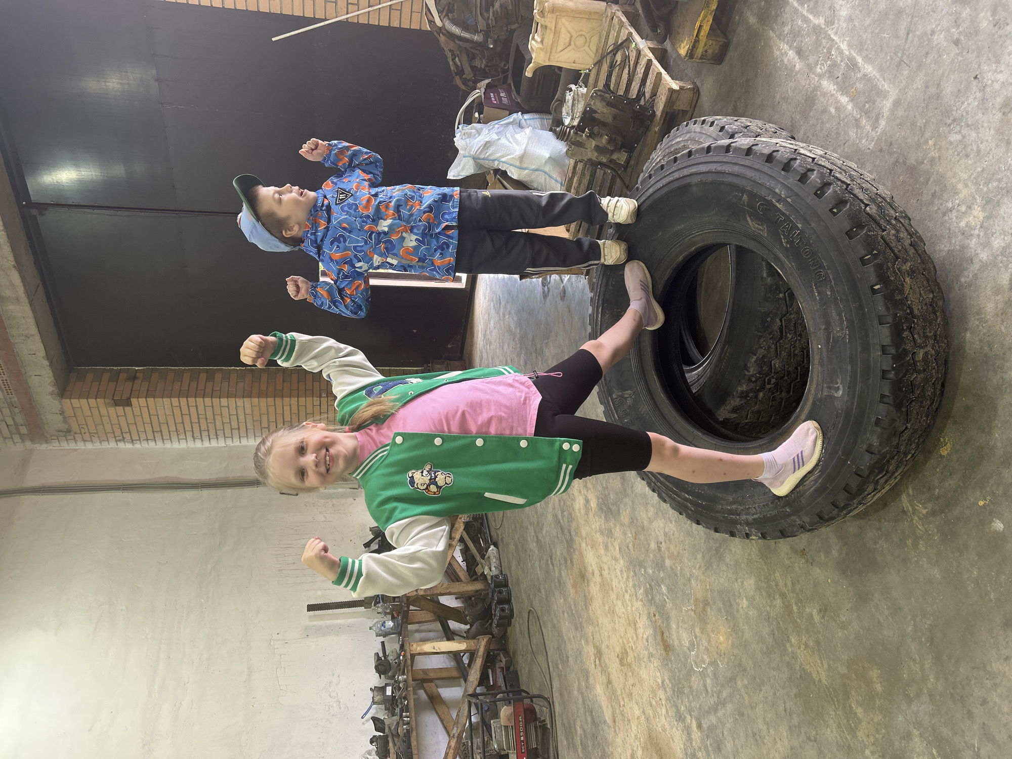 In an interior possibly located in an auto repair shop or storage area in the city of Chaikovsky, Russia, two children pose on and near a large tire. A girl with blonde hair, wearing a pink t-shirt, black shorts, and a green and white varsity jacket, stands with one leg extended onto the tire and both arms flexed in a celebratory gesture. Above her, a boy wearing a blue patterned jacket, black pants, a light blue baseball cap, and sneakers is standing atop the tire, also with his arms raised. The background includes a dark garage door, brick walls, a concrete floor, and various pieces of equipment and tools.