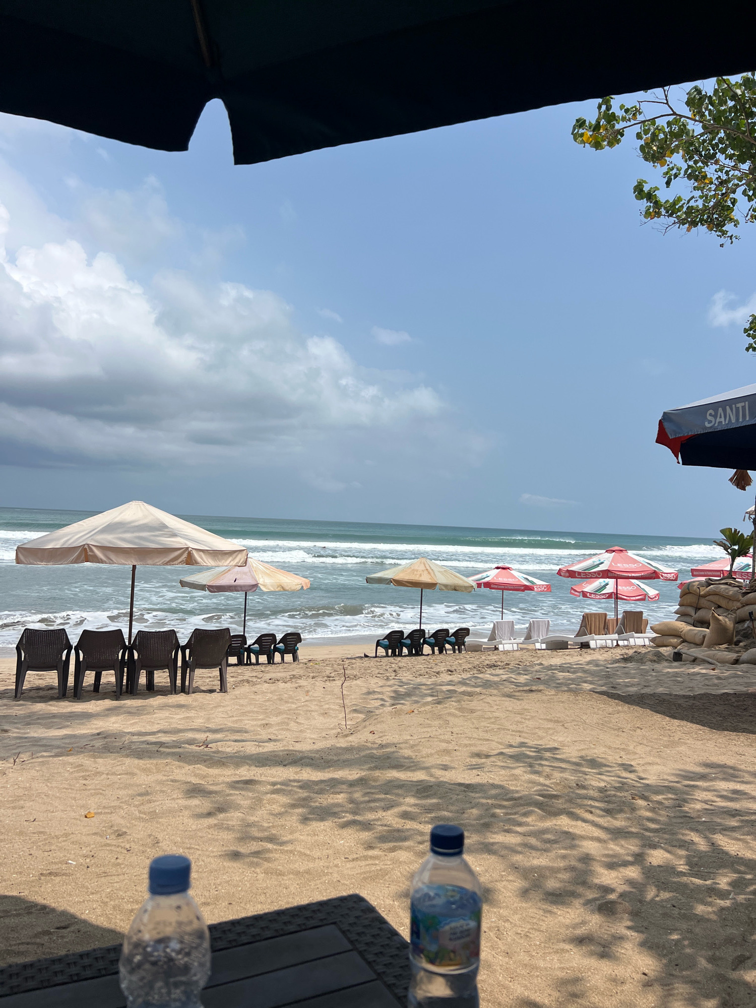 A scene of a beach in Kuta, Indonesia, is depicted. The foreground shows two plastic water bottles resting on a dark, textured surface, possibly a table. Beyond the bottles, the sandy beach stretches towards the ocean. Beach chairs and umbrellas are arranged on the sand, some with people faintly visible relaxing under them. The ocean waves are rolling in, and the sky is partly cloudy with patches of blue. To the right, sandbags are piled up, and a large umbrella with the word "SANTI" is visible. The overall atmosphere is that of a relaxed beach day.
