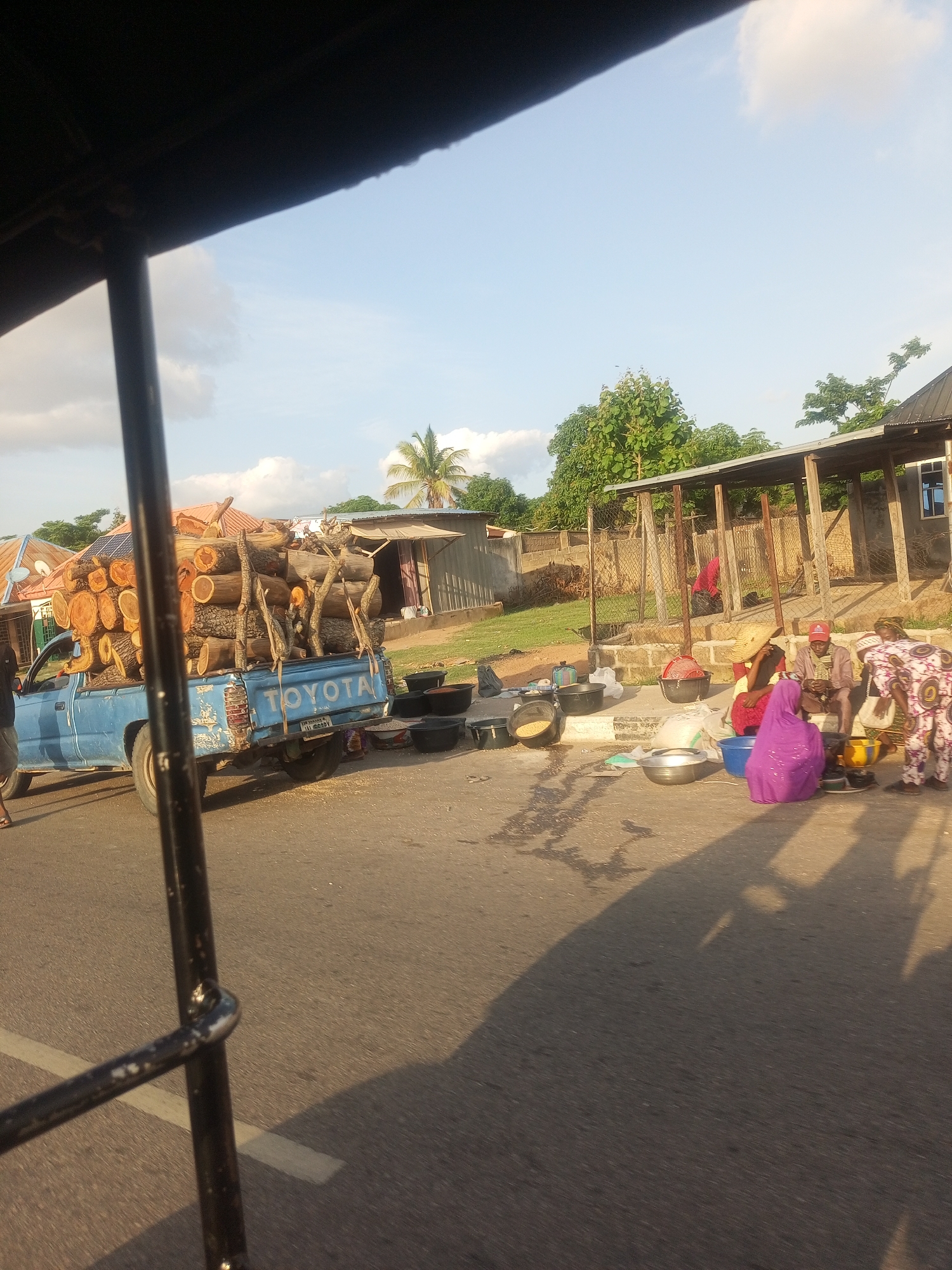 In Jalingo, Nigeria, a light blue pickup truck is parked on a paved road, heavily laden with cut logs. The truck, a Toyota, has "TOYOTA" emblazoned on its tailgate. In the background, several people are gathered near a market stall with a corrugated metal roof. Some individuals are seated, while others stand. Various containers, including large black basins and metal bowls, are present in the scene, some appearing to hold goods. A few individuals are wearing colorful traditional clothing, and one person is wearing a red cap. The sun is low in the sky, casting long shadows, suggesting it is late afternoon or early evening. The weather appears clear with a few scattered clouds visible in the blue sky. The overall scene depicts a marketplace or roadside selling environment.
