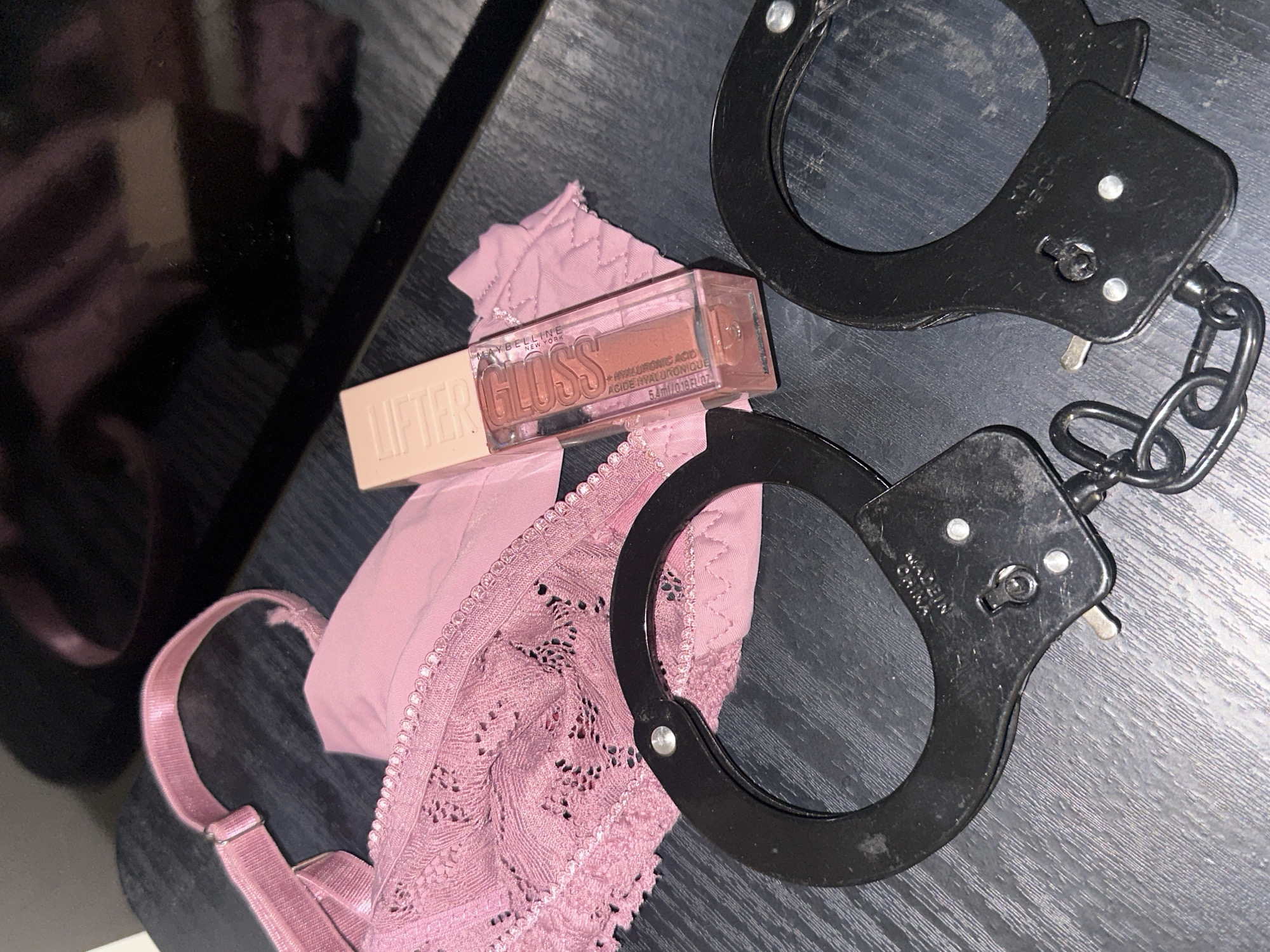 The image displays a still life arrangement of several objects on a dark, textured wooden surface in the city of Sofia, Bulgaria. A pink lace bra is positioned on the left side of the frame, revealing intricate lace patterns on its cup and an adjustable strap with a metallic buckle. Adjacent to the bra, to its right, lies a smaller piece of plain pink fabric, possibly part of an undergarment. Diagonally placed near the center of the image is a clear, rectangular tube of Maybelline New York Lifter Gloss lip gloss, containing a light brown/nude liquid. The tube's cap is beige, and the label indicates the presence of "HYALURONIC ACID." In the foreground, towards the bottom right, are a pair of black metal handcuffs connected by a short chain. One cuff clearly shows partially inverted text, "MADE IN CHINA." The background features a dark, reflective surface with blurred pinkish reflections. No individuals or active interactions are visible within the scene.