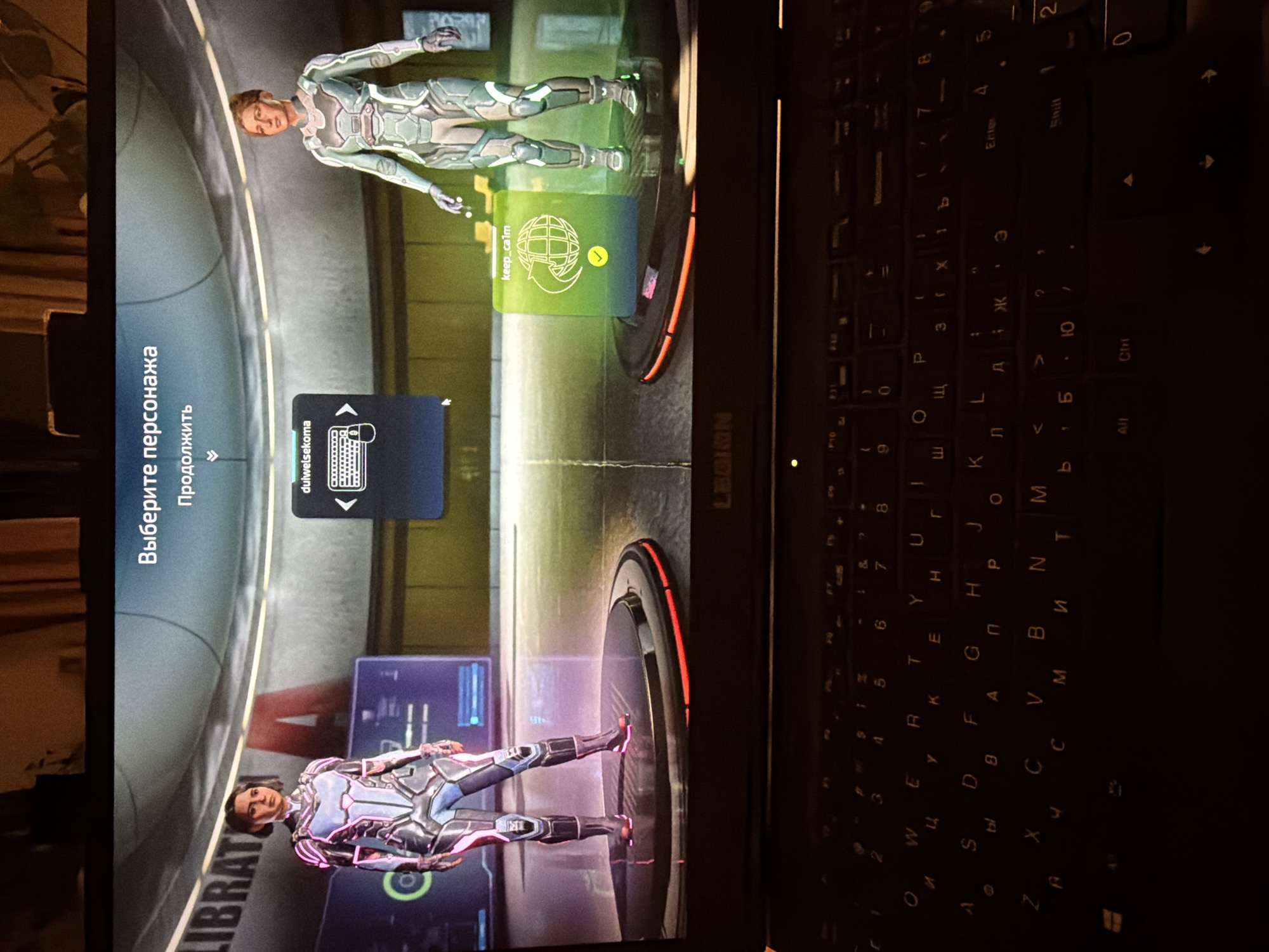 The image shows a laptop screen displaying a video game. Two characters in futuristic suits stand on platforms. Text in Russian appears on the screen, including "Выберите персонажа" (Choose Character). A keyboard, partially visible, is adjacent to the screen. The game appears to be set in a futuristic environment. There is no information about the location in the city of Boreti, Montenegro.