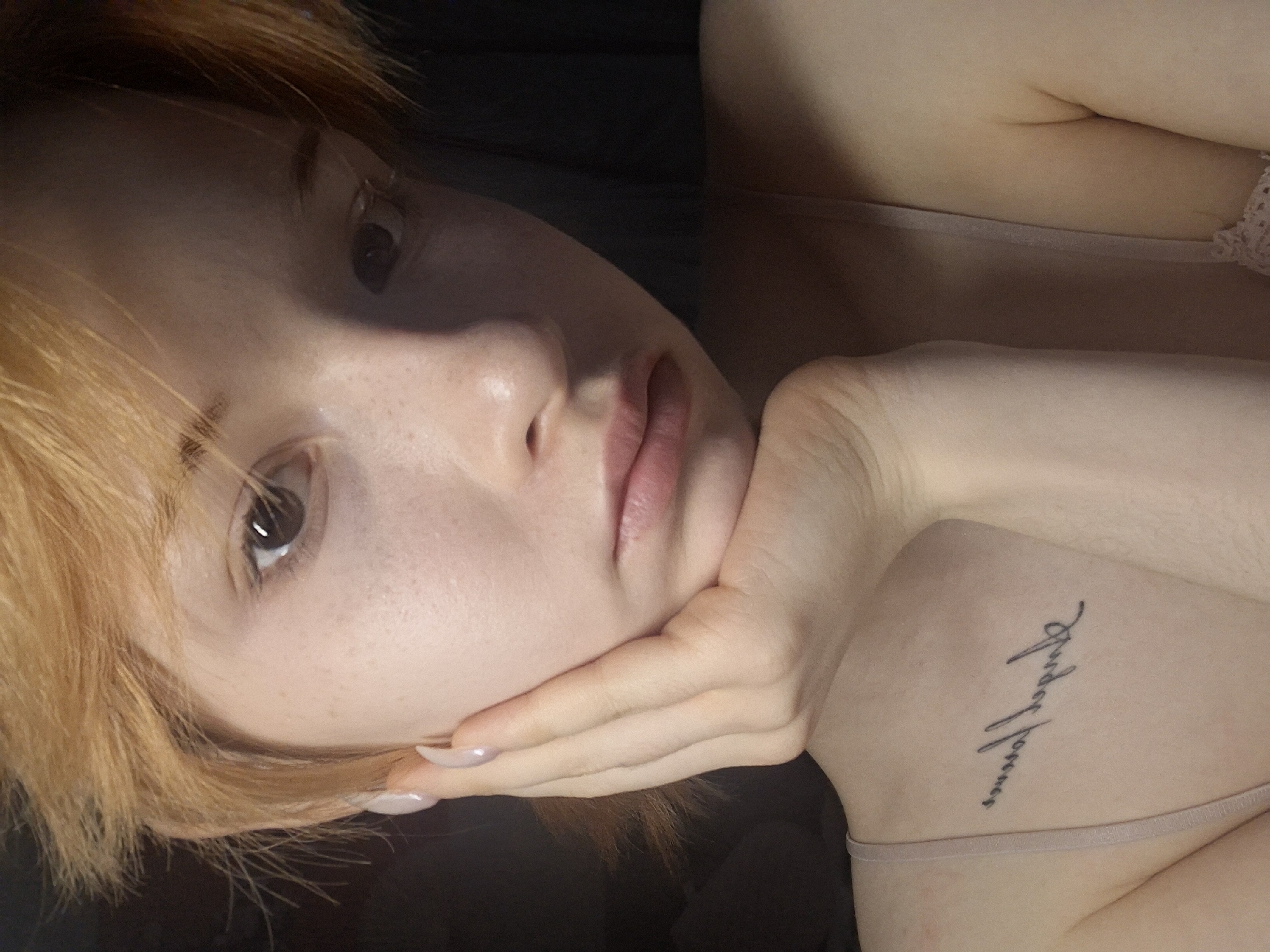A close-up image shows a woman with fair skin and reddish-blonde hair. Her face is turned toward the camera, and her left hand is supporting her chin. She has light freckles and appears to be wearing minimal makeup. A thin strap suggests she is wearing a camisole or similar garment. On her upper shoulder, there is a script tattoo. The background is dark, and the lighting is soft. There is no visible information about a specific location, like the city of Chaikovsky, Russia.