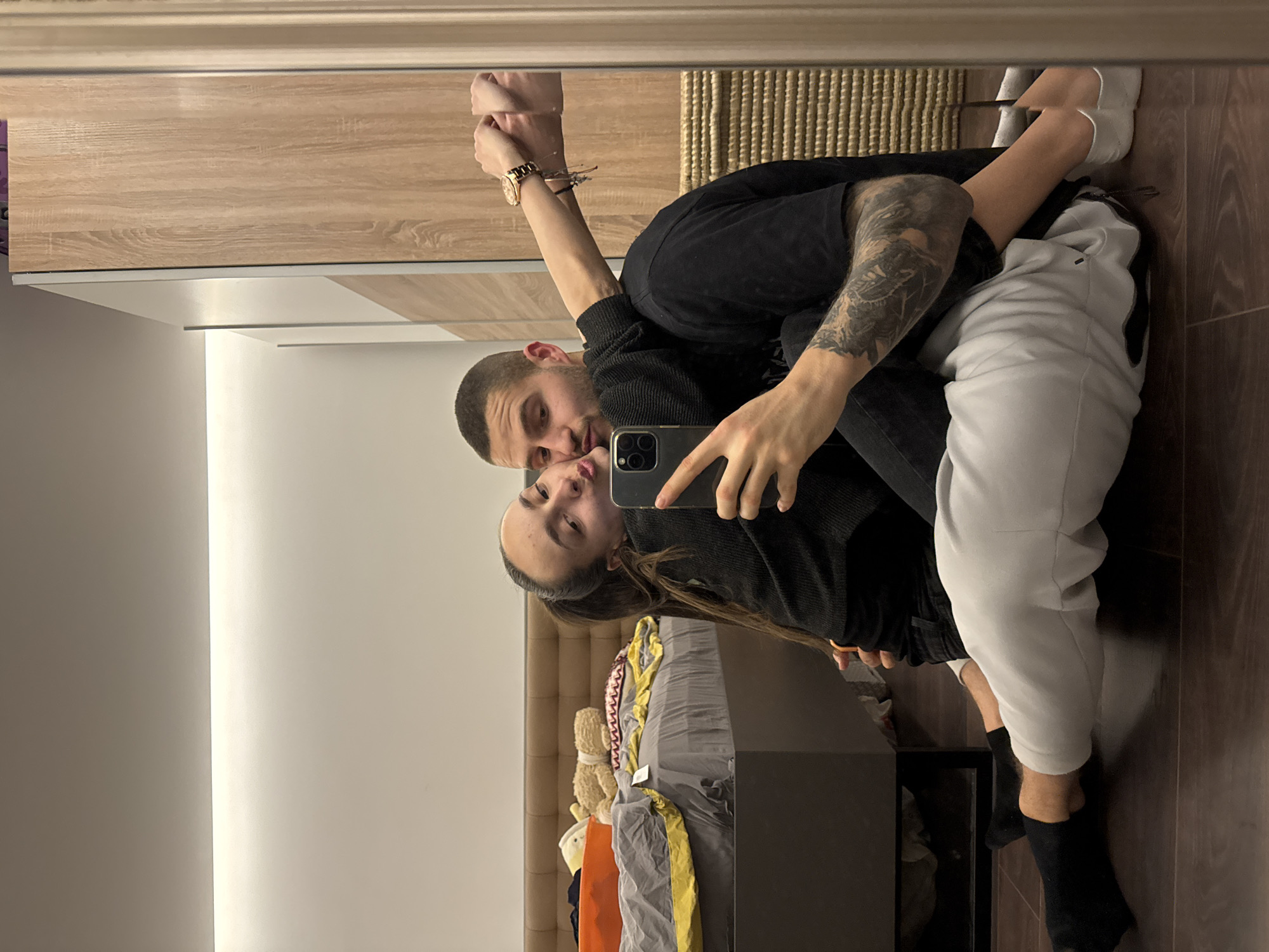 The image displays a mirror selfie of two individuals, a male and a female, seated in an embrace on a dark brown wooden floor within an indoor residential setting in Sofia, Bulgaria. The female, positioned in front of the male and leaning back against him, has dark hair tied in a ponytail and wears a black long-sleeved top and black pants. She holds a dark-colored smartphone in her right hand, capturing the reflection. Her lips are puckered in a kiss gesture directed towards the male, and her left arm is wrapped around his neck. The male, seated behind her, has short dark hair and wears a black t-shirt and light gray sweatpants. His left arm is around the female's waist, and his right hand holds her left wrist, which is adorned with a gold-toned wristwatch and multiple bracelets.
Reflected in the mirror, to the left of the couple, is an unmade bed with a light brown upholstered headboard. The bed is covered with gray and yellow bedding, featuring a patterned pillow, a brown teddy bear, and another plush toy, alongside an orange container. A dark, possibly reflective, bedside table stands next to the bed. To the right, a large wooden wardrobe or cabinet with light wood grain panels and white accents is visible in the reflection. The room features a white ceiling with a linear LED light strip illuminating the wall junction. A portion of a light wood-grained door frame or furniture panel is directly visible on the right edge of the frame, and a woven textured object is partially visible behind the male's lower legs.
