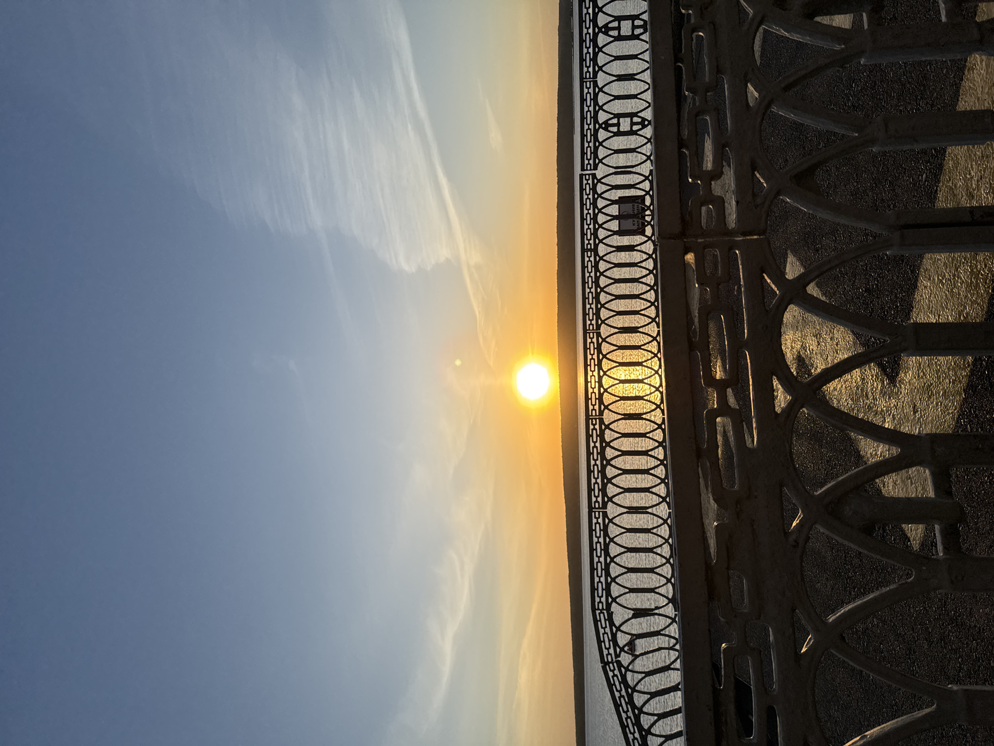 The image depicts a sunset over the Kama River in Naberezhnye Chelny, Russia, as viewed through a decorative iron fence. The sun is positioned in the center of the frame, casting a bright glow across the water and sky. The sky transitions from a clear blue on the left to a warm orange and yellow gradient towards the horizon. Thin, wispy clouds are visible in the sky. The Kama River appears as a dark, reflective surface beneath the sunset. The iron fence occupies the right side of the image, with light reflecting off its surface. The ground behind the fence is dark.