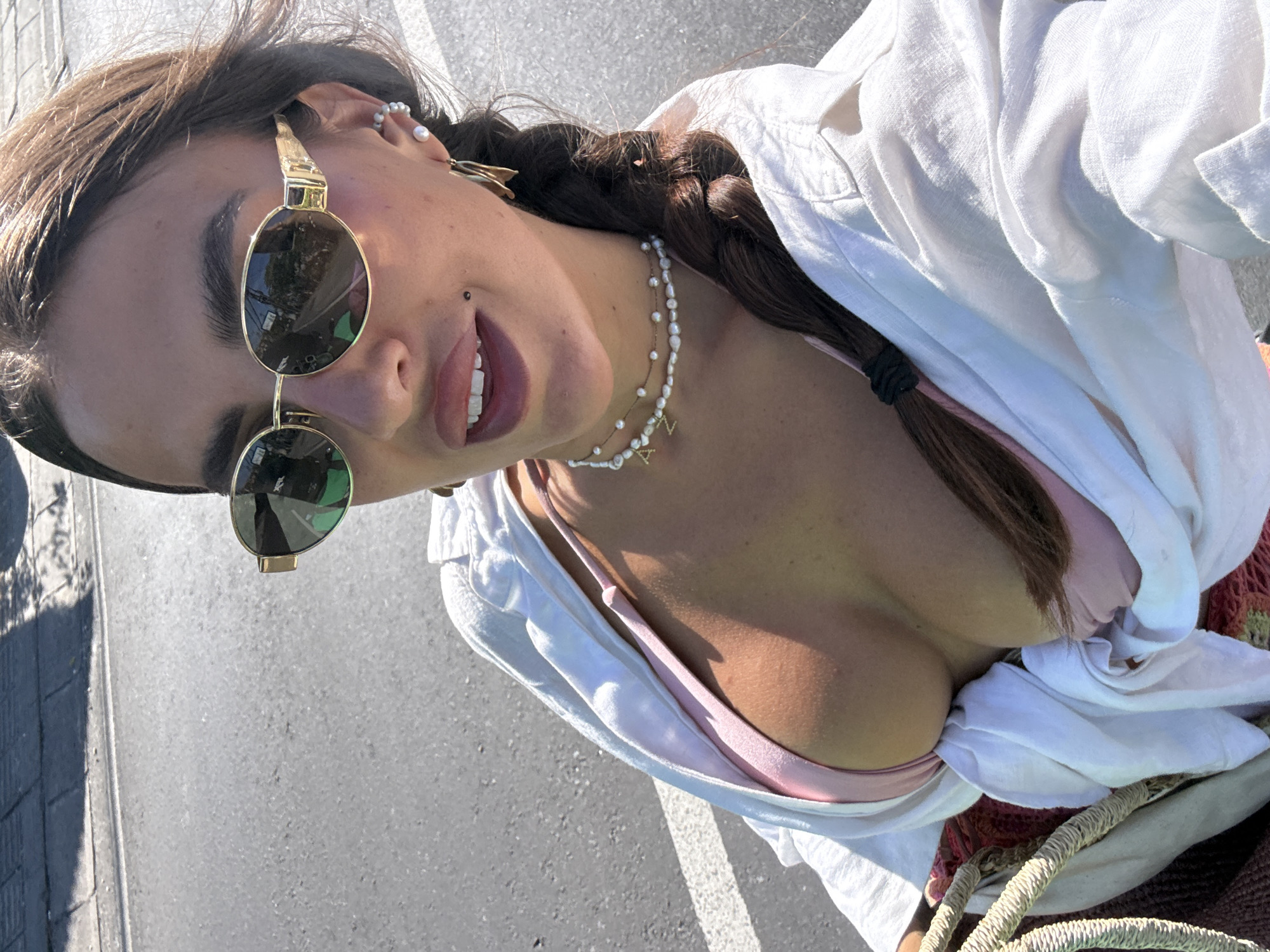 A young woman is captured in a close-up, low-angle, eye-level shot in Kuta Selatan, Indonesia. She wears round sunglasses with a gold frame, a white button-down shirt that is partially open, and a pink top underneath. Her dark hair is styled in a braid. She has a mole near her lip, is wearing pearl earrings, and a necklace with letter charms. The environment appears to be an outdoor area, possibly a parking lot or a street, given the asphalt visible in the background. The sunlight is bright, indicating it is daytime.