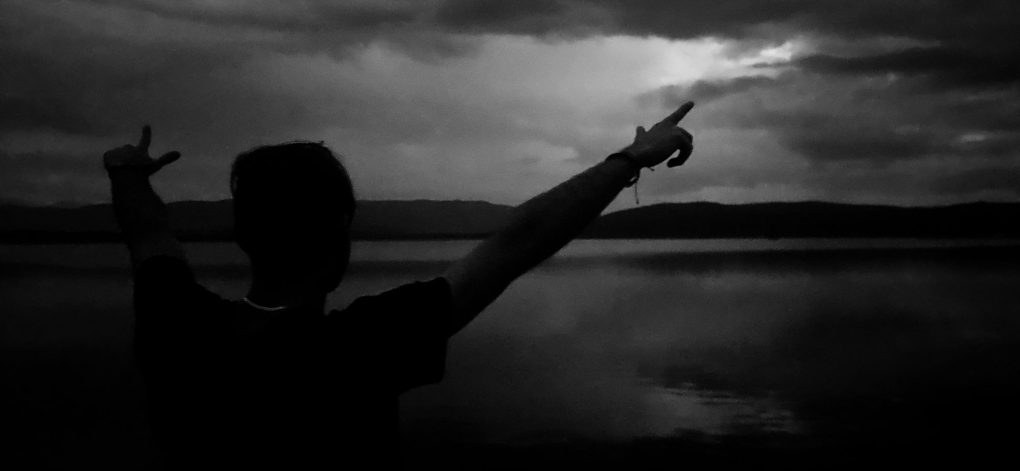 A black and white photograph captures the silhouette of a person standing by a body of water, facing away from the viewer. The individual is positioned in the foreground, with their back to the camera, looking out towards a vast landscape. Their left arm is raised and bent at the elbow, displaying a hand gesture resembling a "shaka" or "hang loose" sign, with the thumb and pinky extended. Simultaneously, their right arm is stretched out and pointed upwards with the index finger, directed towards the cloudy sky. The person appears to be wearing a t-shirt and has a thin necklace visible around their neck.
In the mid-ground, a calm body of water, likely a lake or a large river, stretches horizontally across the frame, reflecting the dim light from above. Beyond the water, a dark line of distant hills or mountains forms the horizon. The sky above is heavily overcast with dark, textured clouds, suggesting either dusk, dawn, or an intensely cloudy day. A slightly brighter patch of sky is discernible towards the upper right, offering a subtle highlight against the predominant gloom.
The image conveys a dramatic and contemplative mood, amplified by the monochrome palette and the stark contrast between the silhouetted figure and the natural surroundings. The gestures of the person could be interpreted as an expression of freedom, a celebratory stance, or an act of pointing towards something unseen. The scene depicts a serene natural environment, typical of areas around Verinsko, Bulgaria. No visible text is present in the image.