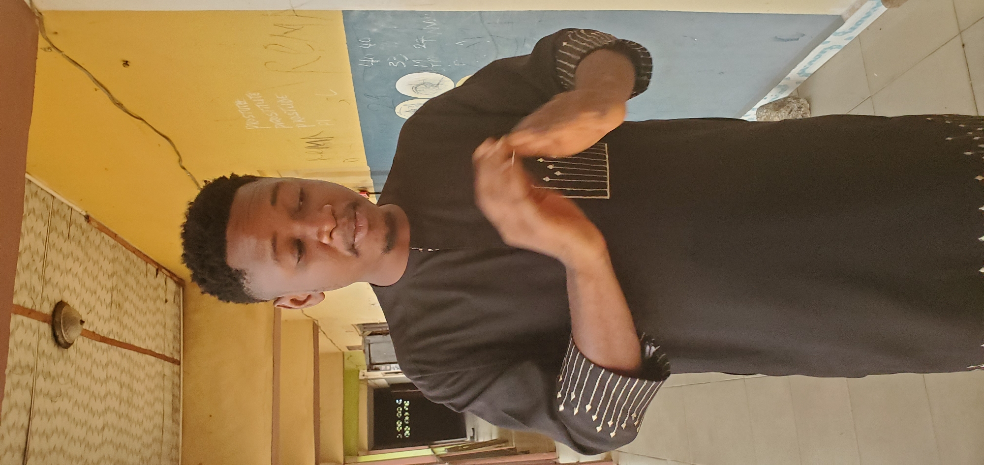 A man is indoors in Mararaba, Nigeria. He is standing in a hallway in front of a wall that has a blue and yellow background with white writing on it. The man has dark hair and dark skin. He is wearing a dark shirt with light-colored stripes on the cuffs. He is holding his hands together in front of him.