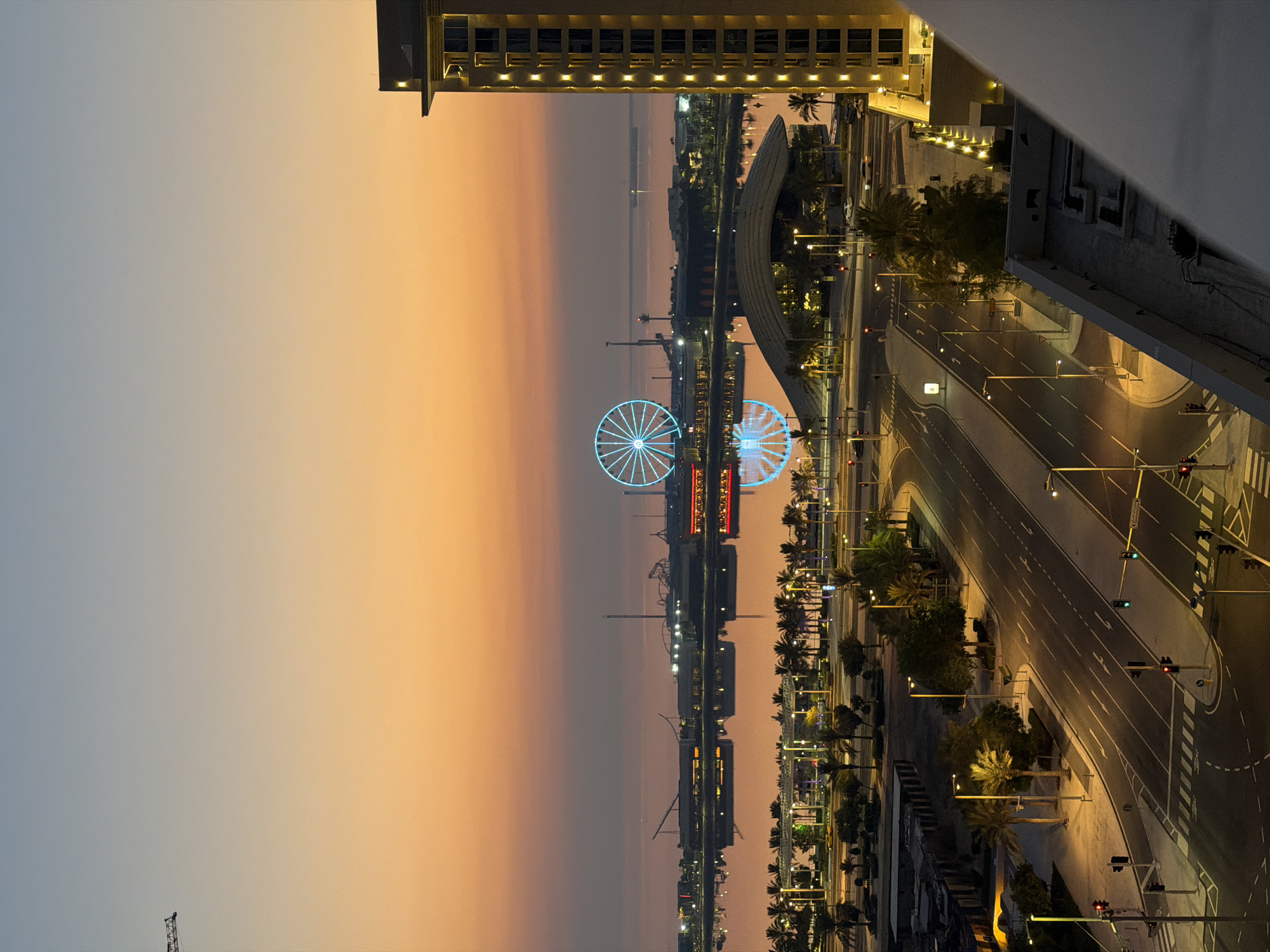 This is a vertical, medium shot of a city scene at dusk. The sky is a gradient of soft blues and oranges, with the sun having just set. In the distance, a large Ferris wheel glows with blue lights, its reflection shimmering on the calm water below. Across the water, a cityscape with various structures, including roller coasters and other amusement park rides, is visible. The foreground features a multi-lane road with streetlights casting a warm glow. Palm trees line the sides of the road and the waterfront. A modern, tall building with illuminated windows stands prominently on the right side of the image, partially obscuring the view of the city. The scene appears to be from a high vantage point, possibly a balcony or a tall building, looking down and across the city.