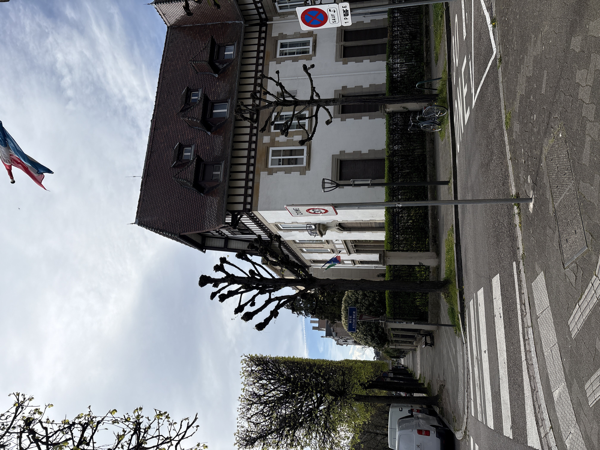 The image depicts a street scene in Strasbourg, France. The primary focus is on a pedestrian crossing with white stripes on the asphalt road. A building with a traditional Alsatian architectural style is visible on the left side of the street. The building has a brown tiled roof with dormer windows and white walls. A French flag is visible, hanging from a pole. A road sign indicating parking restrictions and a speed limit of 30 are present near the building. The street is lined with trees, some of which are pollarded. A sign for "Rue Stoeffer" is also visible. Parked cars are present on the right side of the street.
