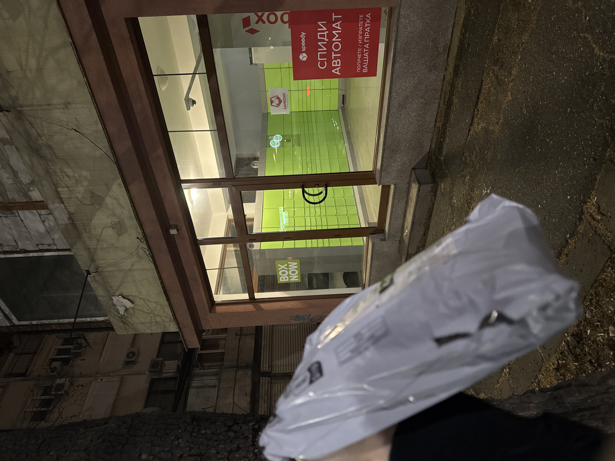 A person is holding a package in front of a Speedy Automate location in Plovdiv, Bulgaria. The storefront is visible, displaying the Speedy logo and the words "СПИДИ АВТОМАТ". Inside the store, the automatic lockers are colored in bright green. The exterior of the building is weathered. The street and building exterior are dark, suggesting it is evening or night.