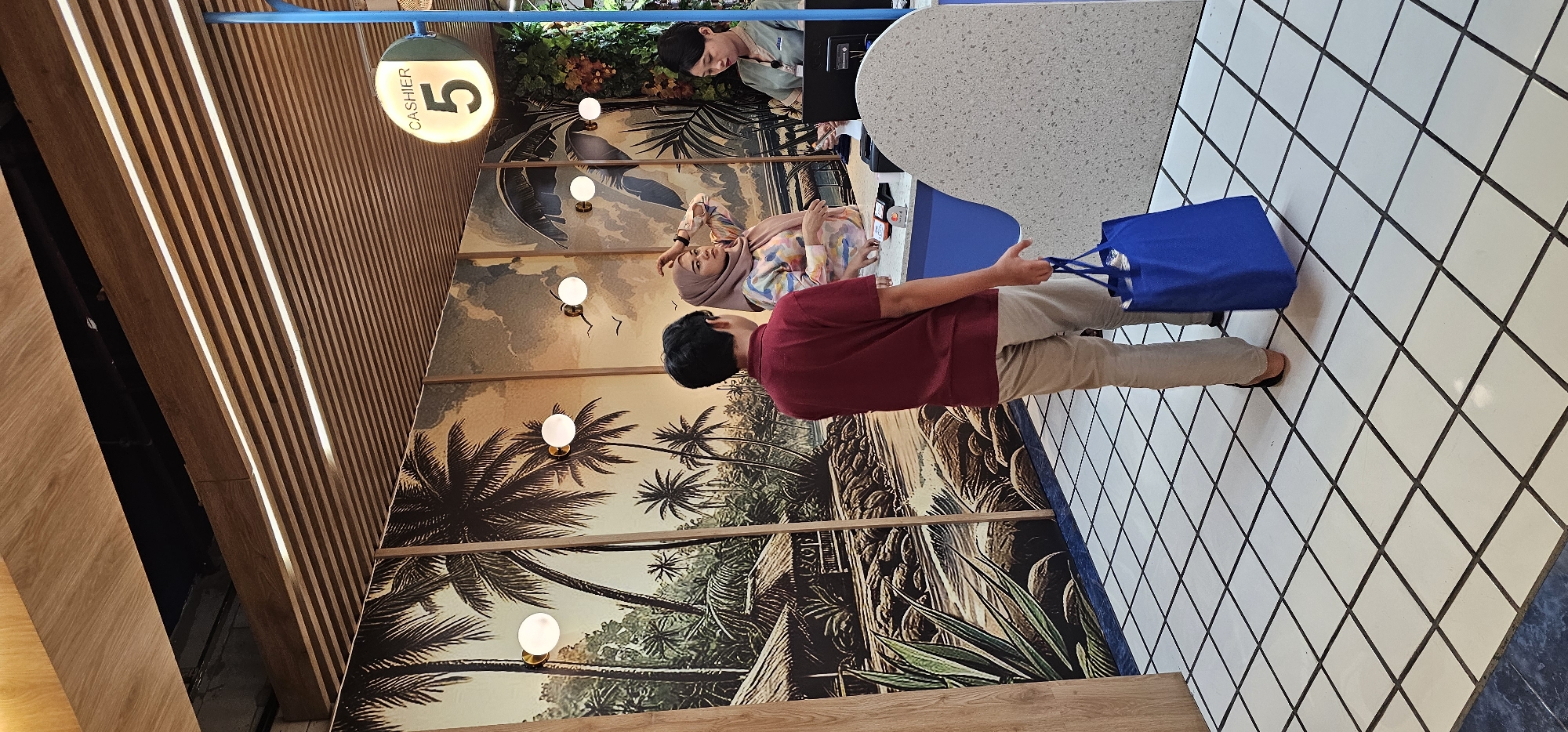 A brightly lit interior space features a tropical-themed mural. Two individuals, a cashier and a customer wearing a headscarf are at the counter. Another customer, facing away from the viewer, is carrying a blue shopping bag. A sign indicating "Cashier 5" is visible. The setting appears to be in Denpasar, Indonesia. The floor is tiled in a white and black grid pattern. The wall behind the counter has a detailed mural depicting palm trees and a river scene, illuminated by globe-shaped lights.