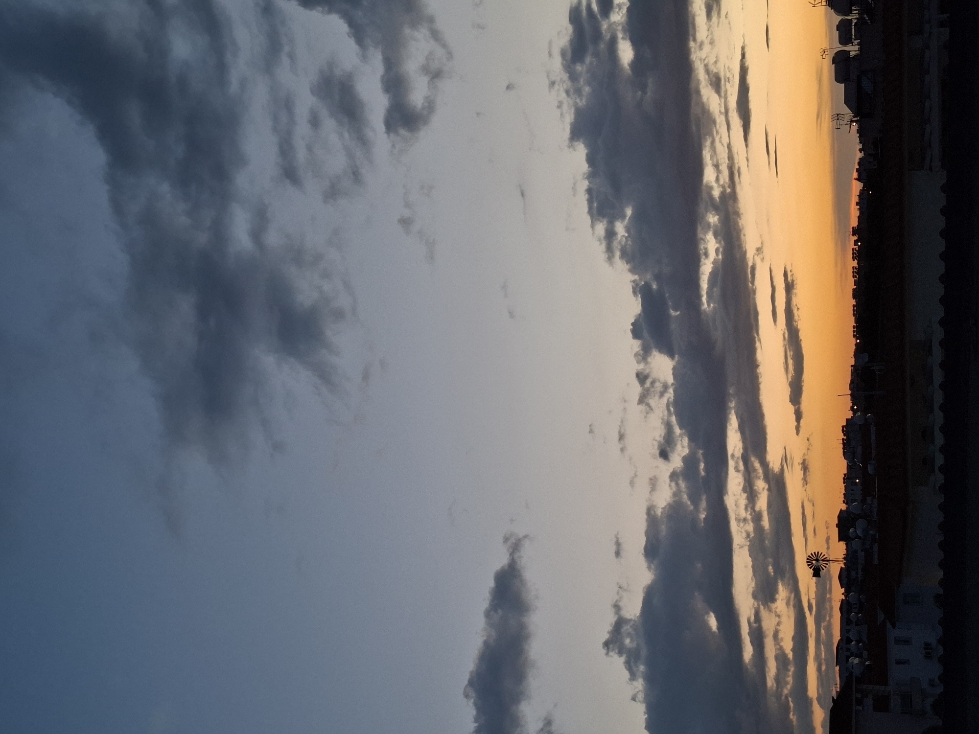 The image shows an orange sunset viewed from Paralimni, Cyprus. A line of buildings is silhouetted against the sunset's orange glow with a small windmill visible in the city skyline. The sky is filled with clouds of varying shades of grey. The darker clouds give way to the blue sky behind them.