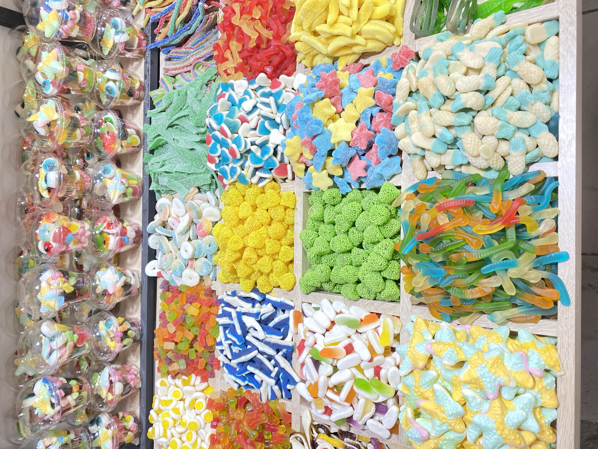 A colorful display of assorted gummy candies is presented in clear plastic cups stacked on shelves, with bulk quantities of various gummy candies arranged in wooden dividers on a table below. The candies include gummy bears, sour belts, fruit shapes, and worm-shaped gummies in a wide spectrum of colors such as red, yellow, green, blue, orange, and white. The backdrop appears to be an indoor setting, possibly a candy store or market stall in Makhachkala, Russia, with a shallow depth of field that blurs the background.