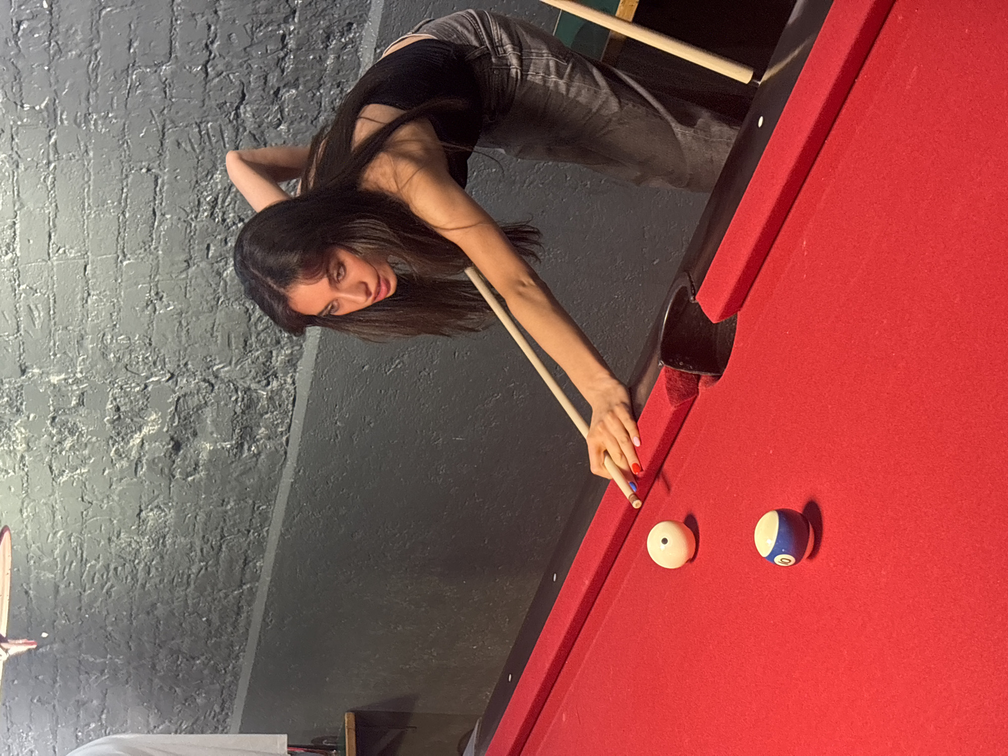A young woman leans over a red pool table, aiming with a cue stick at a white cue ball and a blue striped ball. The scene is indoors, with a textured gray wall in the background. The woman has long dark hair, wears a black top and jeans, and has manicured nails. The pool table has a dark trim, and a pocket is visible. The location is not discernible from the image.
