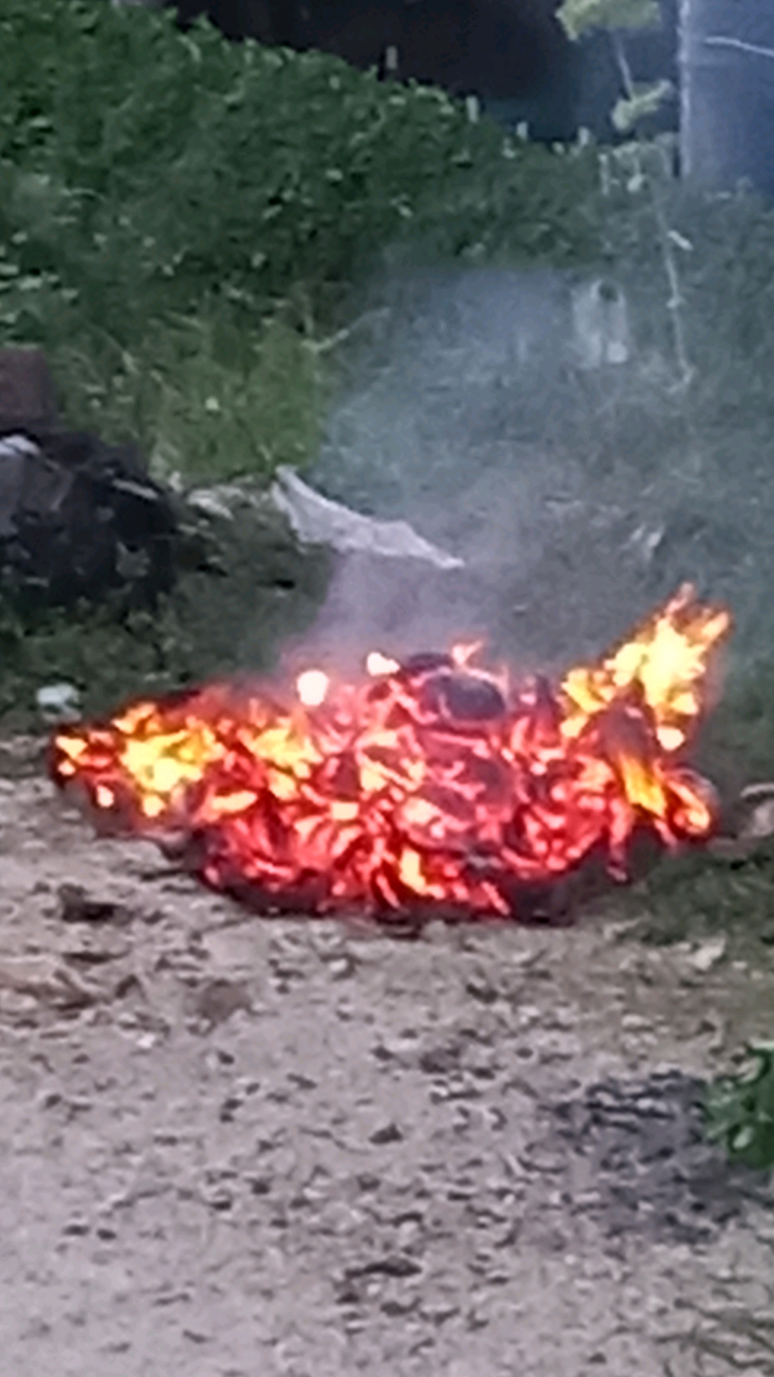 A pile of burning material, appearing to be leaves and possibly wood, is situated on a dirt path in an outdoor environment. The flames are a bright orange and yellow, with some smoke rising from the fire. In the background, there is a grassy area with some foliage. The scene is set in the Ko Pha-ngan District of Thailand.