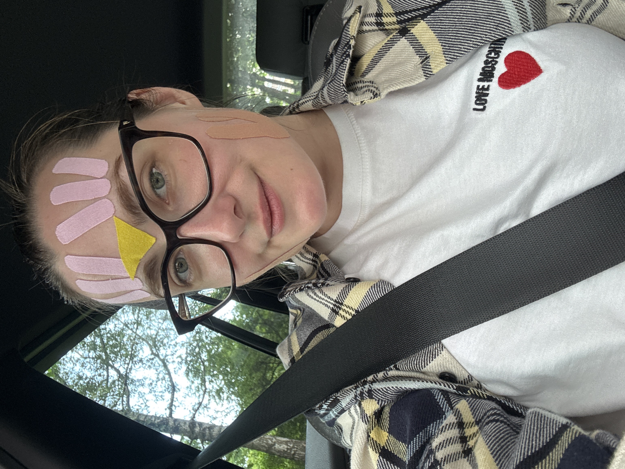 A person with glasses and tape on their forehead and cheek is sitting in a car. They are wearing a white t-shirt with "LOVE MOSCHINO" and a red heart printed on it, and a plaid shirt over it. A seatbelt is visible across their chest. The background shows trees and foliage, suggesting an outdoor environment. The image does not contain enough information to determine the specific location within the city of Riga, Latvia.