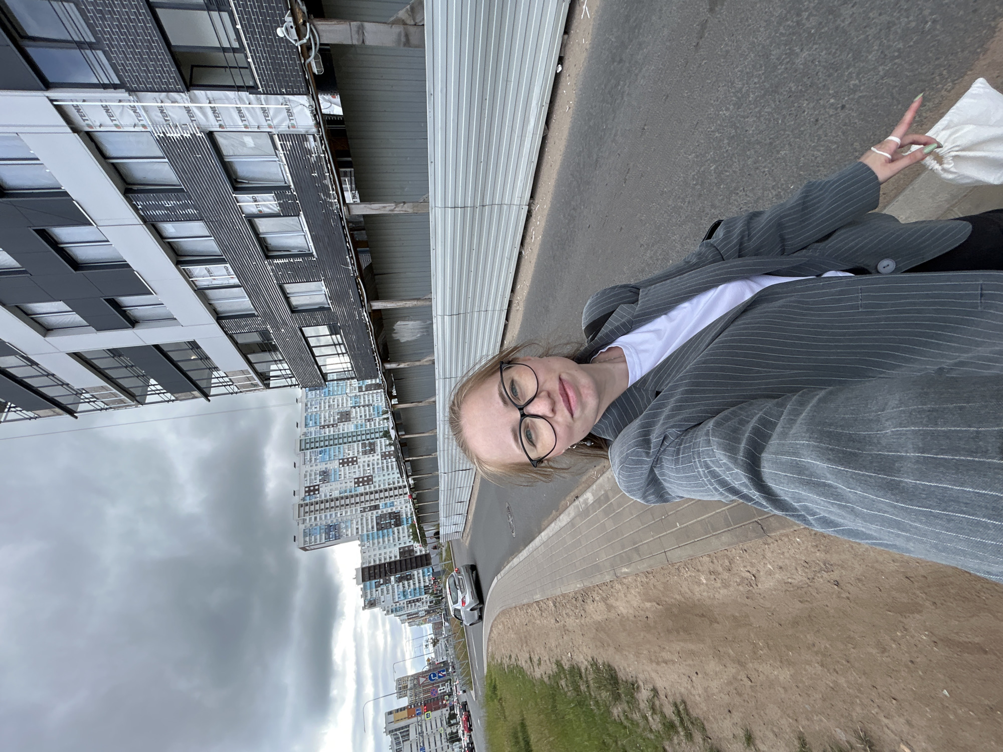 This media file is a portrait-oriented selfie taken outdoors. The primary subject is a young woman with blonde hair, wearing glasses and a pinstriped grey blazer over a white t-shirt. She is holding a white bag in her left hand and making a peace sign with her right hand, wearing several rings and having green nail polish. She appears to be smiling or smirking.
The setting is an urban environment in Saint Petersburg, Russia, characterized by modern, multi-story residential buildings. To the left of the woman, a new, unfinished building is visible, with exposed construction materials and scaffolding. Further in the background, a row of completed, tall residential buildings with many windows dominates the skyline. A street with cars and traffic signs runs alongside the woman, with a paved sidewalk and a grassy verge. The sky is overcast and cloudy, suggesting a possibly cool or dull day. The time of day is not definitively clear but appears to be daytime. The overall mood seems casual and perhaps a bit posed. There is no visible text in the image.