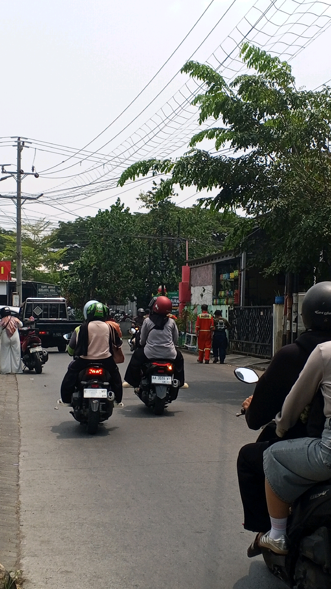 The image depicts a street scene in Semarang, Indonesia, with several motorcycles and people on the road. In the foreground, two people are riding a motorcycle, with the rider wearing a black helmet and the passenger in a light gray top and jeans. To the left, another motorcycle carries two people, with the rider wearing a green helmet and a light brown and gray top, and the passenger in a light brown top. Further down the road, a person in a white and patterned headscarf and white pants is walking away from the viewer, beside a black pickup truck.
On the right side of the street, two men in orange jumpsuits are standing near a building. One man is looking towards the street, while the other is facing away. Another person in dark clothing is walking past them. Buildings with various signs and decorations line the street, and trees with green foliage are visible. Overhead, numerous electrical wires crisscross the sky, some of them supported by poles. The overall atmosphere is that of a busy, urban environment during daylight.