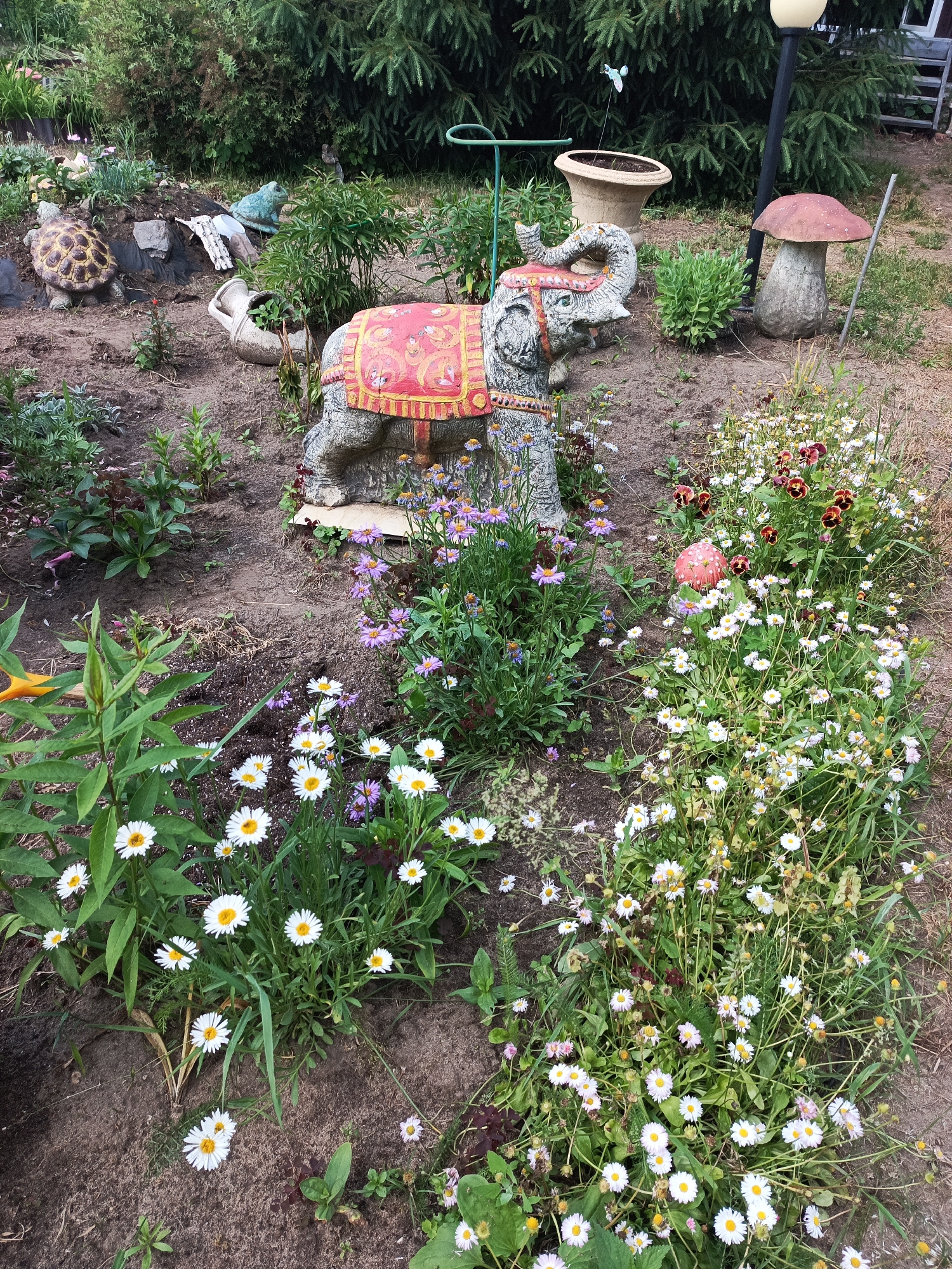 A garden scene in Novoe, Russia, features an elephant statue adorned with a red and gold blanket, positioned among various plants and garden ornaments. To the left, a turtle statue sits near a large terracotta pot and a ceramic frog. In the background, evergreen trees and a lamppost are visible. A large mushroom-shaped ornament stands to the right of the elephant statue. The ground is covered with dirt, dry grass, and an assortment of flowering plants, including white and purple daisies and dark red pansies.