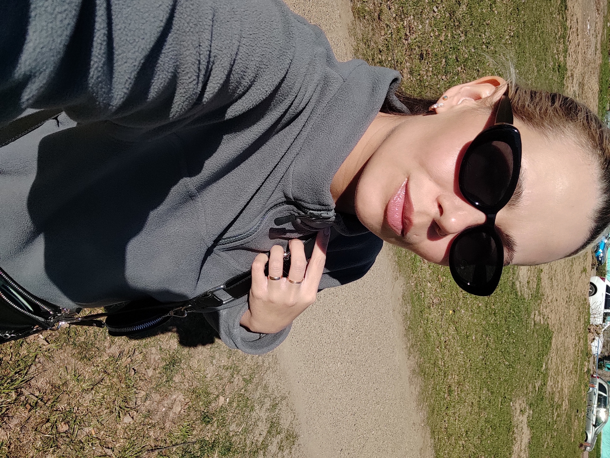 A woman stands in a grassy area with a dirt path in what appears to be Chaikovsky, Russia, as evidenced by visible vehicles in the background. She is wearing a gray fleece jacket and black sunglasses, holding a black handbag. She has two rings on her left hand and a piercing in her ear. The scene is captured from a slightly angled, elevated perspective, possibly a selfie. The lighting suggests it is daytime.