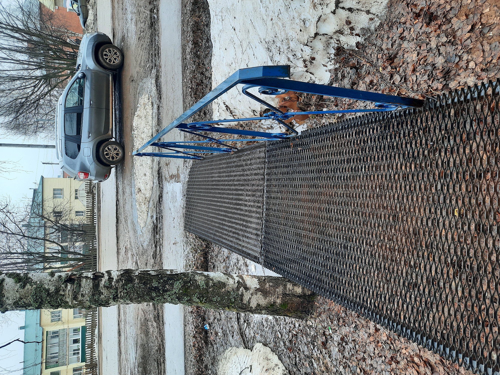 The scene depicts a slushy, possibly snow-melting environment, likely a street or path, within the town of Krasnogorskoye, Russia. A grey hatchback car is parked adjacent to a raised metal grid walkway with a blue railing. A birch tree trunk is positioned near the walkway. The ground is covered in dead leaves and melting snow. Buildings are visible in the background. No people are visible in the frame.