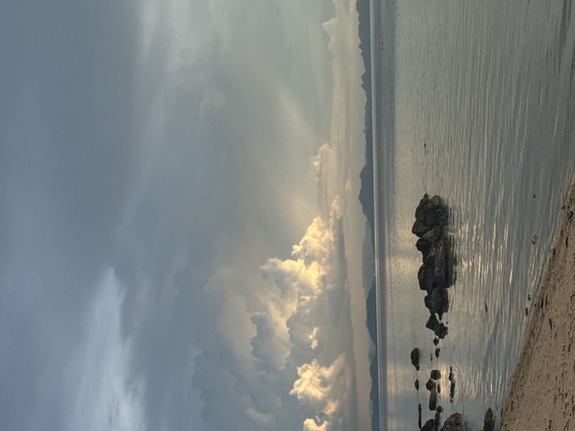 The image depicts a serene coastal scene. The foreground features a sandy beach transitioning into the calm, reflective waters of the sea. Several dark, wet rocks are visible along the shoreline and partially submerged in the water, some forming a small cluster. In the distance, a hazy silhouette of mountains lines the horizon. The sky is dramatic, with large, fluffy cumulus clouds tinged with yellow and white from sunlight breaking through, juxtaposed against a backdrop of overcast, grey clouds. Rays of sunlight are visible piercing through the clouds, creating a celestial effect. The overall impression is one of natural beauty and tranquility. This scene is located in Baan Pang Ka, Thailand.
