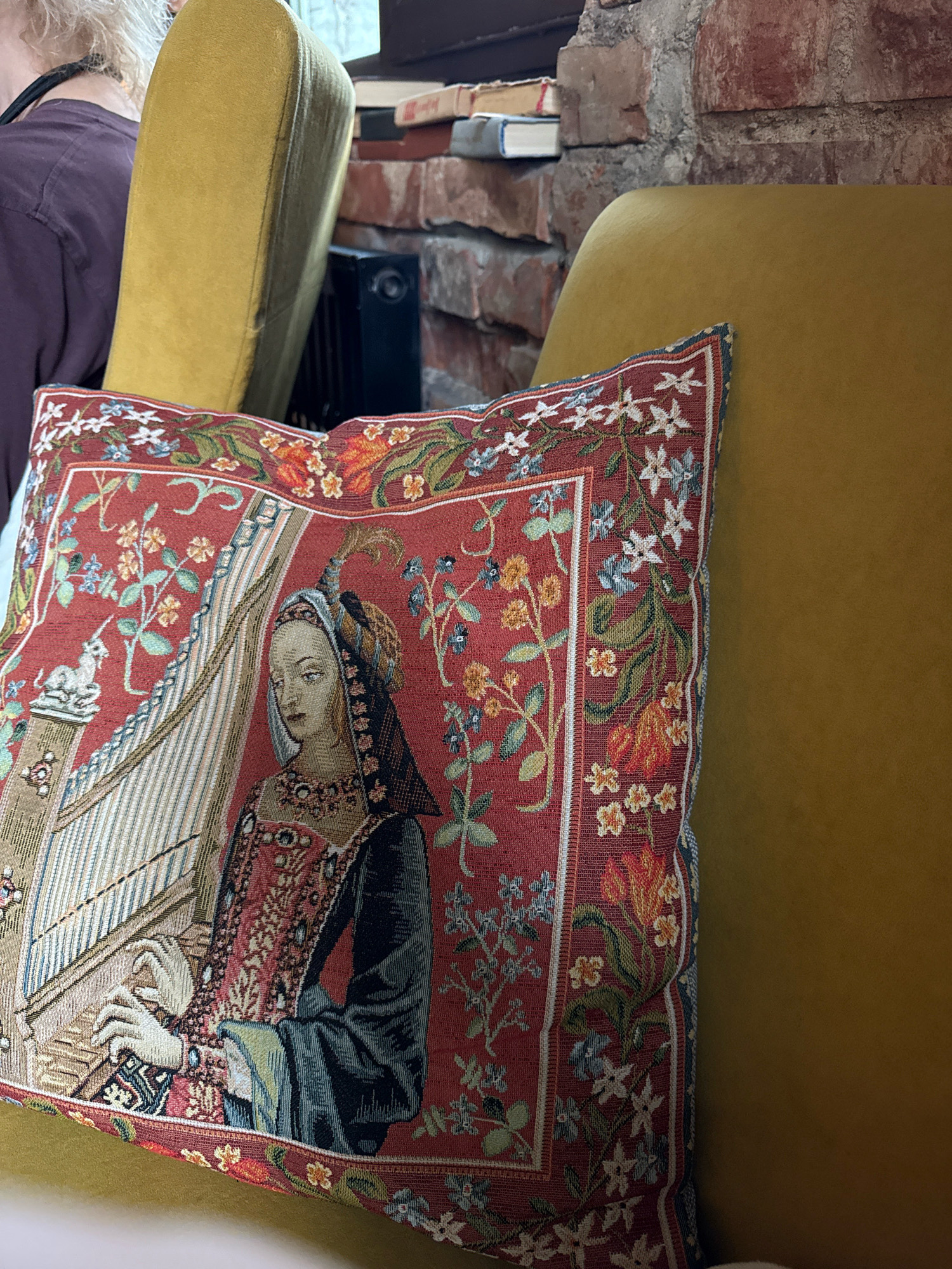 A close-up, eye-level shot shows a tapestry pillow with a medieval scene of a woman playing a harp. The pillow is red with a floral border and depicts the woman in elaborate clothing, a detailed harp, and a white unicorn. The pillow rests on a mustard-yellow armchair. In the background, a brick wall and a stack of books are visible. A portion of a person wearing a dark purple shirt is visible on the left side of the image. The scene appears to be indoors, likely in a domestic setting or a cafe. There is no information to indicate this image was taken in Kaliningrad, Russia.