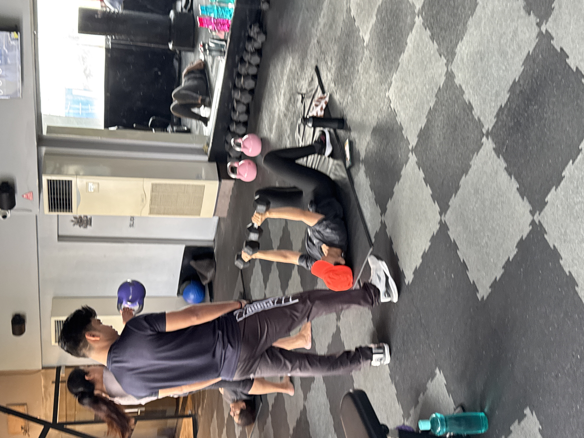 **Scene:**
The image shows an indoor gym setting, likely in Kolkata, India, given the presence of Indian individuals. The gym has a gray and black diamond-patterned floor and a wall with a mirror and air conditioning unit.
**People:**
Several people are present in the gym, engaging in physical activity.
One person is lying on their back on a mat, lifting dumbbells. They are wearing black leggings and a dark shirt. A red cap is lying nearby.
Another person is standing and lifting a kettlebell.
Another person is standing and appears to be working out.
Another person is lying on the floor in the background
**Objects:**
Dumbbells of varying sizes are lined up against the wall.
Kettlebells of pink and blue colours are also present.
Water bottles are scattered on the floor.
A gym mat is laid out on the floor.
A towel and phone are lying on the floor.
**Actions:**
People are engaged in various forms of exercise, including weight lifting and strength training.
**Interactions:**
The individuals seem to be working out independently.
**Environment:**
The gym appears to be a functional space with standard exercise equipment.