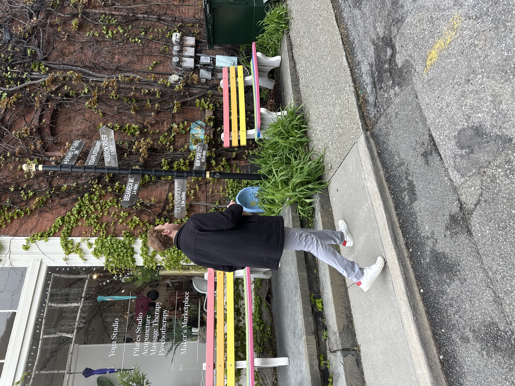 The image depicts a daytime street scene in the Village of Cold Spring, United States of America, with an overcast sky. A man, viewed from his left side, walks along a concrete sidewalk. He is wearing a black jacket, light grey pants, and white sneakers, with light brown curly hair and a beard, and appears to be looking upwards.
To his left is a rustic brick building facade, extensively covered in green ivy and dry vines. Multiple utility boxes are mounted on the wall. In front of the building, two decorative wooden benches are visible, painted in vibrant stripes of pink, yellow, blue, and purple. A blue bucket labeled "GOZILLA" rests on the ground near the benches.
A tall black pole stands in front of the building, adorned with several wooden directional signs pointing to various local and distant destinations. The visible text on these signs includes: "ASCEND STUDIO 50ft," "YOGA PILATES BARRE," "HUDSON RIVER 1,168ft," "BREAKNECK MT. 2.4 miles," "GLYNWOOD 3.94 miles," and "NEW YORK CITY 45.54 miles."
The storefront window of the building displays text, likely advertising services within: "Yoga Studio," "Pilates Studio," "Acupuncture," "Massage Therapy," "Psychotherapy," and "Maker's Marketplace," along with "FL1," "FL2," and "FL3" indicating floors. The sidewalk transitions into an asphalt road on the right, featuring a faint yellow line. The scene suggests a tranquil, small-town atmosphere.