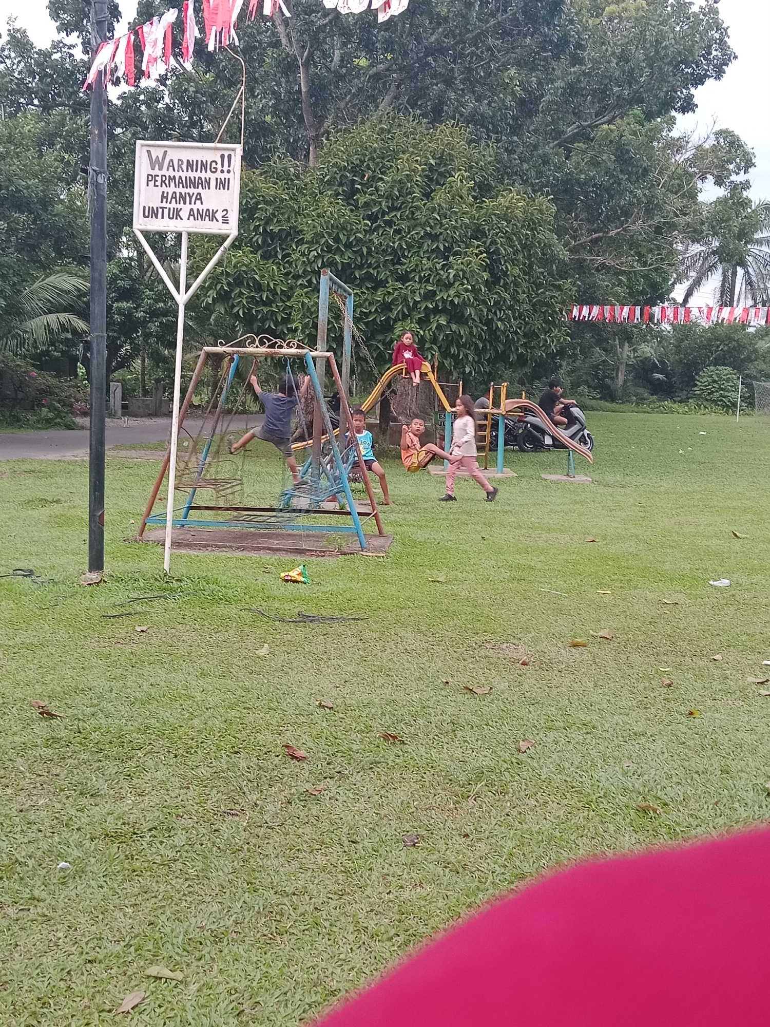 In the City of Binjai, Indonesia, a children's playground is depicted. The playground features a swing set, a slide, and other play structures, all situated on a grassy area. Several children are actively playing on the equipment. A sign is visible, indicating that the playground is intended for children. The background consists of trees and foliage, suggesting a park or recreational area. Decorative Indonesian flags are strung across poles, adding to the festive atmosphere. The weather appears to be overcast.