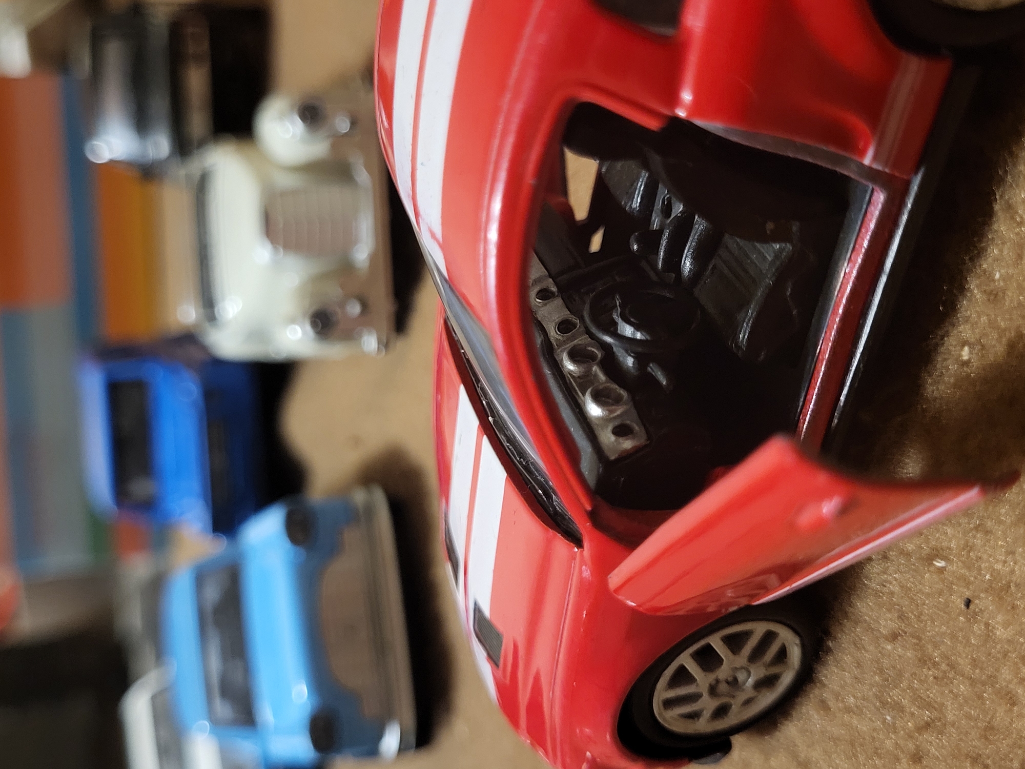 The image shows a close-up view of a red and white toy car, specifically the front portion with the hood open, revealing a detailed engine compartment. To the left of the red toy car, there are several other toy cars, blurred and stacked. The background appears to be a textured surface, possibly carpet or a similar material. The setting and subject matter do not indicate any specific location in Yerevan, Armenia, as it seems to be a collection of toys in a domestic environment.