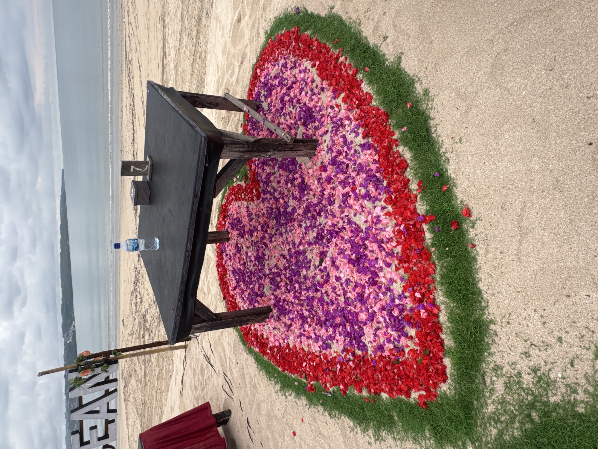 The scene is a beach setting, presumably in Kuta, Indonesia, given the request. A low black table with wooden legs is set on the sand. On the table are a small wooden sign with the number "7" on it, a small woven container, and a bottle of water. The table is situated in the center of a large heart shape made of layered flower petals; the outer layer is red, the middle is pink, and the inner portion is purple. Green grass outlines the heart shape. In the background, the ocean meets the shoreline under a cloudy sky. In the lower left corner, part of a white wooden sign and the corner of a red bench are visible.