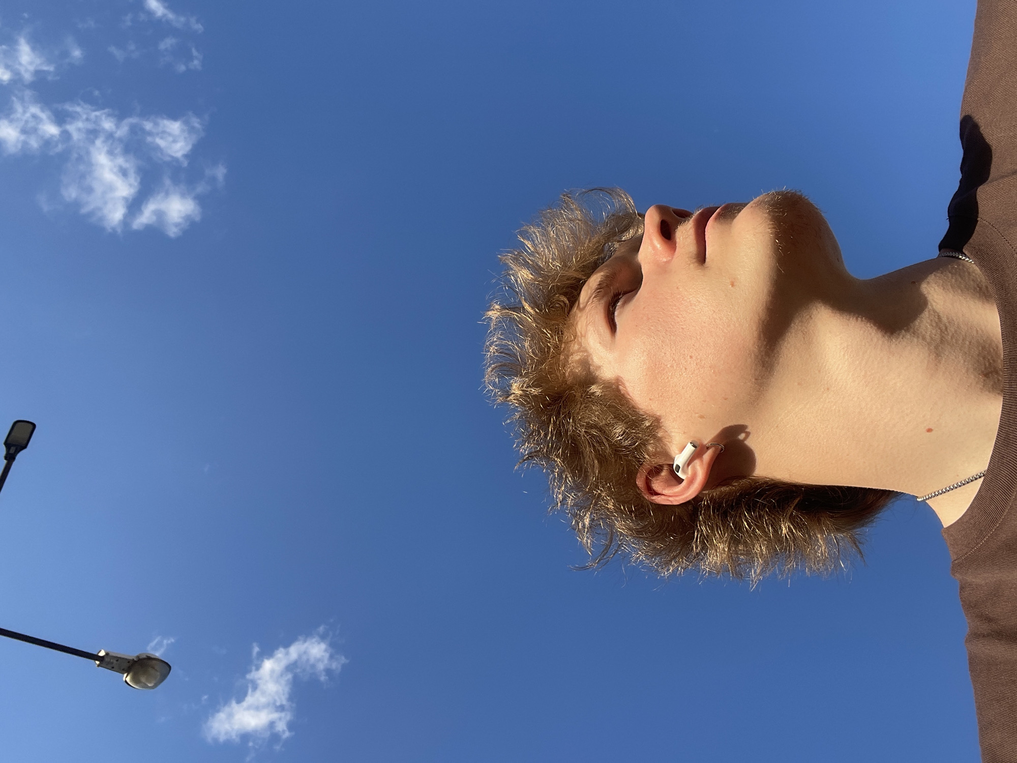 A low-angle view of a person looking up at the sky. The sky is a clear, bright blue with a few scattered white clouds. A street lamp is visible in the lower left corner of the frame. The person has light skin, light brown hair, and is wearing a brown shirt. An earbud is visible in the person's left ear. The location cannot be determined from the image.