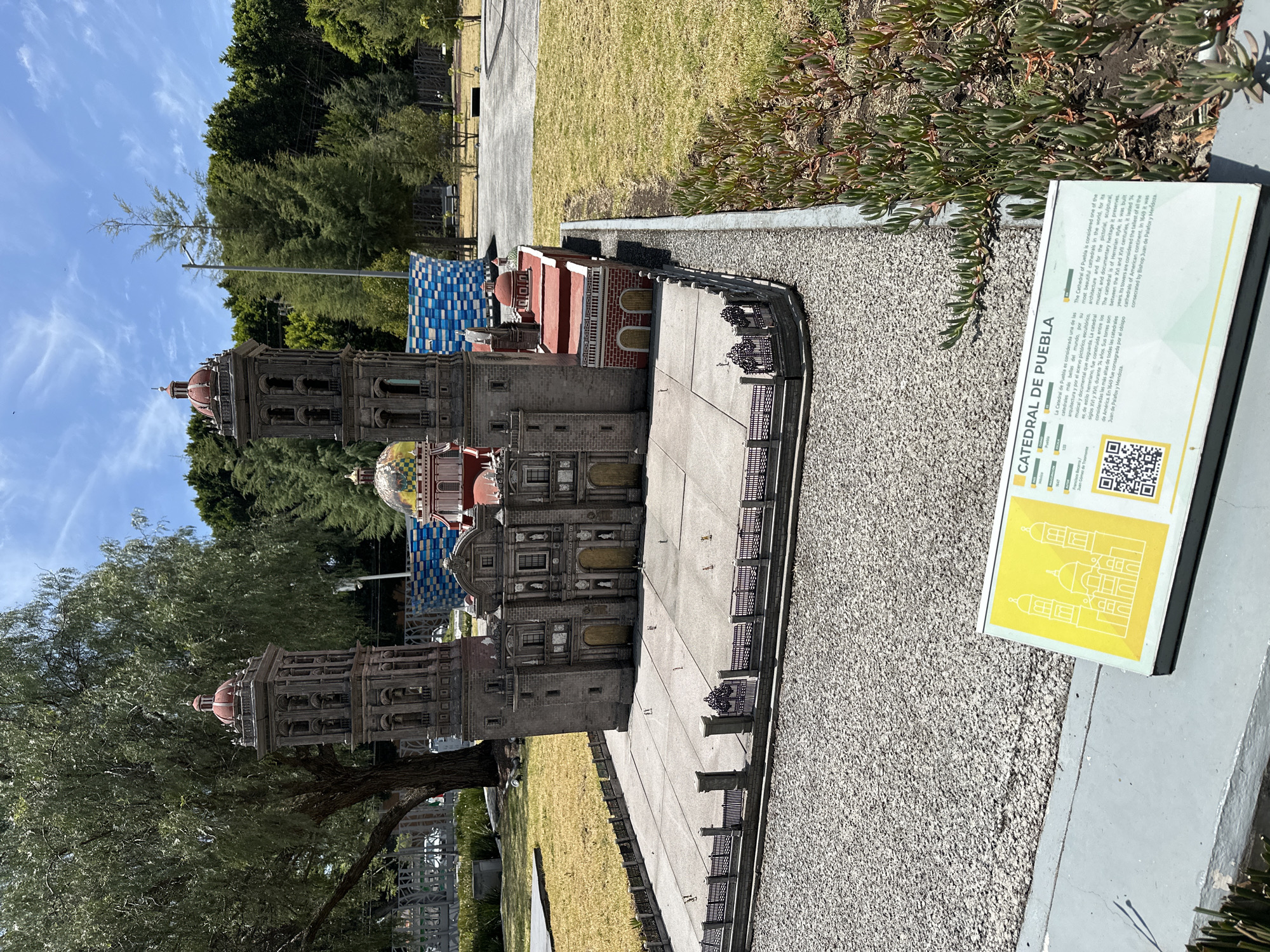 The image shows a miniature model of the Catedral de Puebla, located in Puebla, Mexico. The model is displayed outdoors, with a sign providing information about the cathedral. The sky is clear with some trees and bushes surrounding the model. The model has gray stone walls with red and blue tiled roofs.