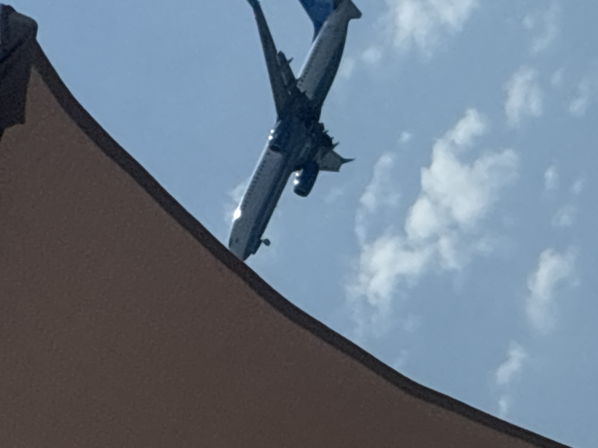 A large, commercial airplane with blue and white markings is captured in mid-flight against a clear blue sky with scattered white clouds. The underside of the airplane is visible, revealing its landing gear. In the foreground, a portion of a large, brown awning or canopy occupies the left side of the frame, partially obscuring the view of the sky. The image appears to be taken from ground level, looking upwards. The location in the city of Sirius, Russia, is not discernible from the visual information provided.