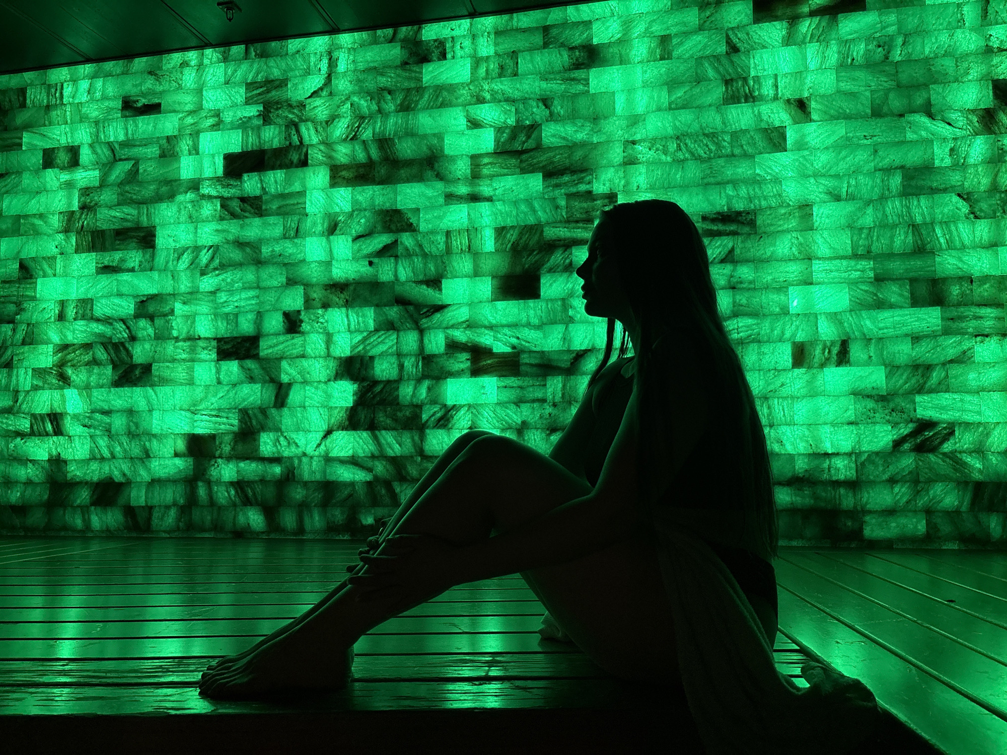 A person sits in silhouette against a brightly lit green wall. The wall appears to be made of rectangular bricks or tiles. The person is seated on what appears to be a wooden floor or platform, also illuminated in green light. The location is a room in Yekaterinburg, Russia.