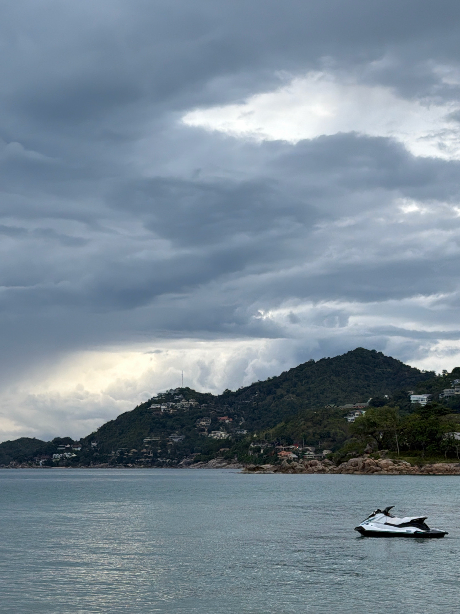 A jet ski floats on calm, grey water in the foreground, its white and black body visible. Behind it, a rocky shoreline is dotted with green trees and houses, leading up to a steep, verdant hill. The hill is covered in trees and more houses, with a prominent communication tower near its summit. The sky above is filled with dramatic, heavy grey clouds, with patches of bright light peeking through in the distance, suggesting a storm. The scene is set in Ko Samui District, Thailand. There are no people visible in the image.